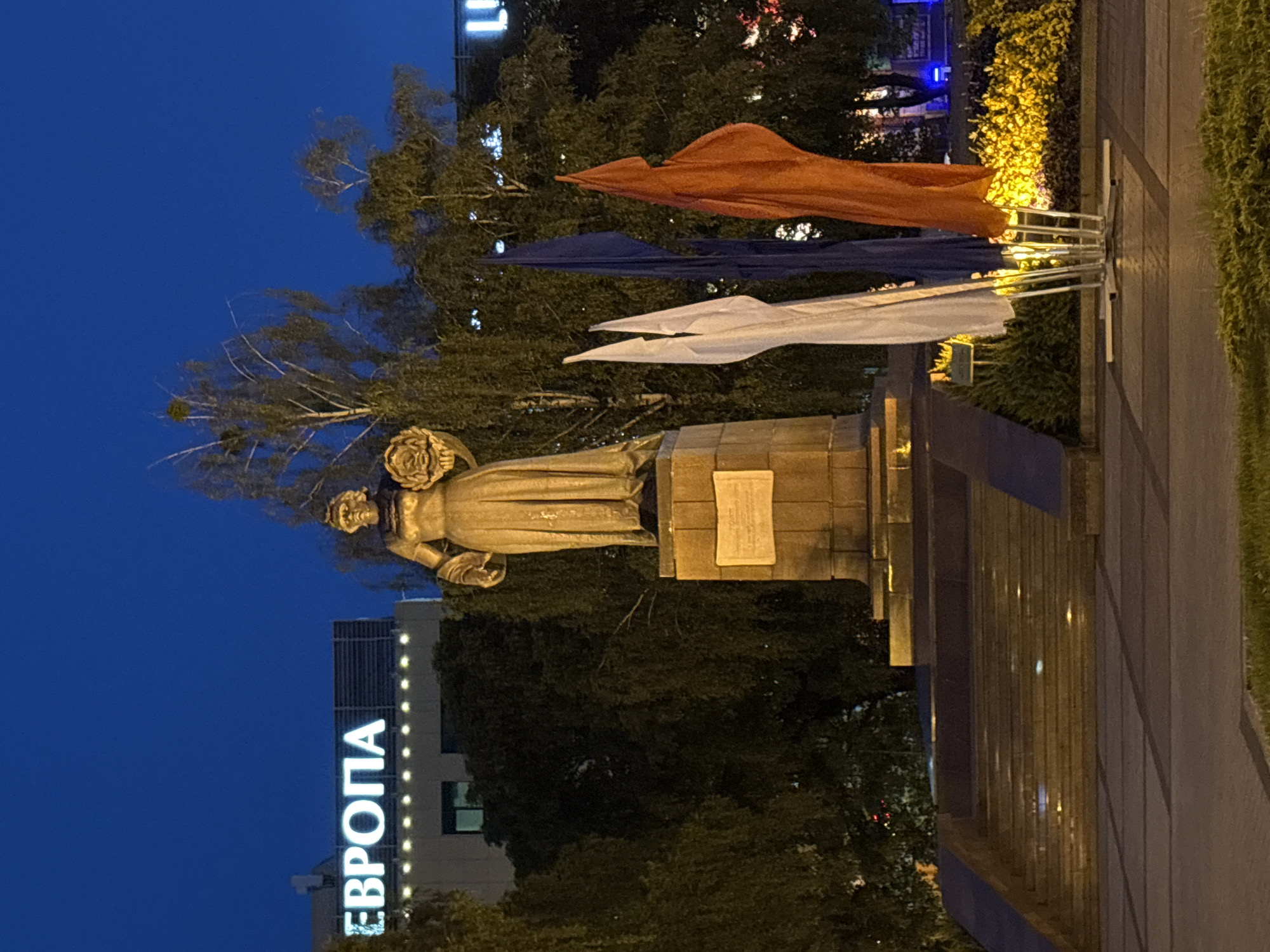 The image captures an urban scene at twilight or night, likely in Kaliningrad, Russia. The central focus is a golden-hued statue of a person, possibly a woman, standing on a pedestal. The figure appears to be holding a bouquet of flowers or a wreath in one arm.
To the right of the statue, three flags or banners are displayed vertically on poles: an orange one at the top, a dark blue one in the middle, and a white one at the bottom. The background is dominated by lush, dark trees and foliage, some illuminated by ambient lights.
On the left side, a modern building is visible, featuring a large, brightly lit sign in Cyrillic script that reads "ЕВРОПА" (EVROPA), translating to "Europe." The building also has vertical light strips. The sky is a deep, clear blue, typical of twilight or early night, with artificial lighting casting a serene glow on the scene. The foreground on the right shows a paved pathway or plaza, bordered by neatly trimmed and illuminated green hedges or bushes. No human activity is explicitly visible, creating a peaceful and illuminated nocturnal view of the monument and its surroundings.
