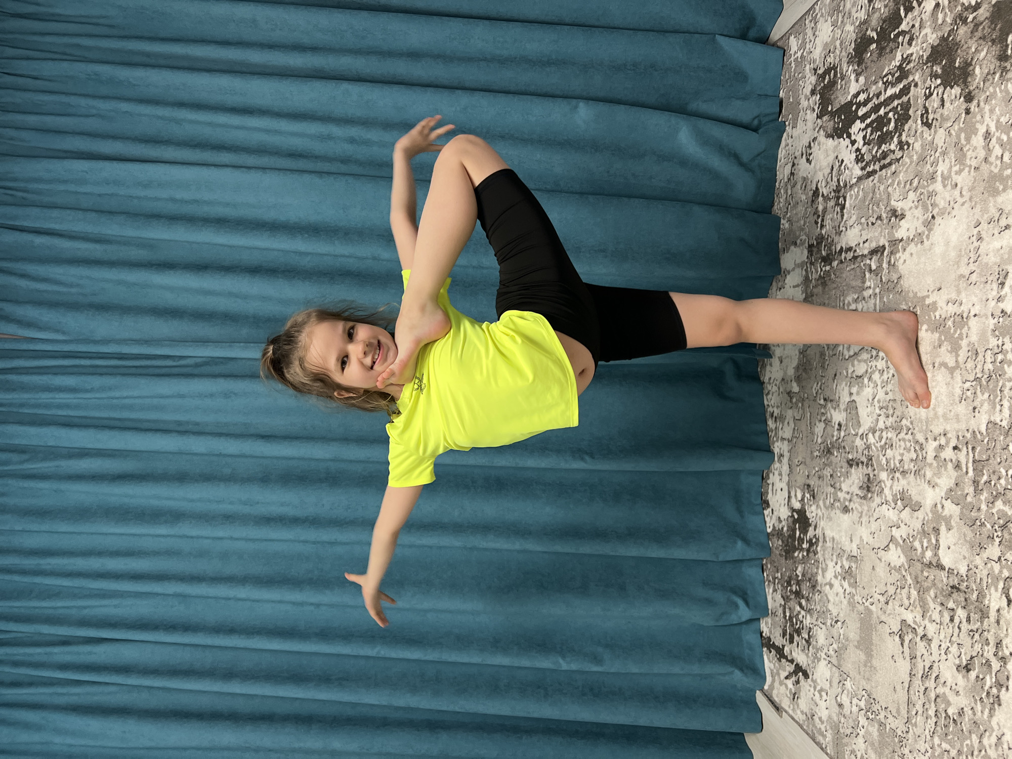 A young girl in a yellow t-shirt and black shorts is captured in a gymnastic pose, with one leg extended straight to the right and the other bent backward, her foot held near her head. Her hair is pulled back, and she is smiling at the camera. The background is a blue curtain, and the floor is covered with a grey and white patterned rug. The picture was taken indoors in Chaikovsky, Russia.