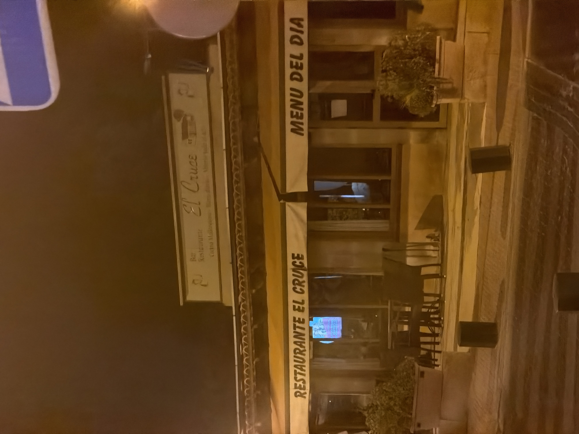 The image shows the exterior of a restaurant named "RESTAURANTE EL CRUCE" at night. The restaurant has a yellow awning with the name in bold black letters. To the right of the awning, there is another sign that reads "MENU DEL DIA". The building is lit from within, and the windows reflect the interior lighting. Outside the restaurant, there are potted plants and outdoor seating arrangements, including tables and chairs. A sign above the entrance reads "Bar Restaurante El Cruce Cocina Mallorquina". The scene is located in the city of Calvià, Spain. The overall impression is a typical Spanish restaurant at night, with a warm and inviting atmosphere.
