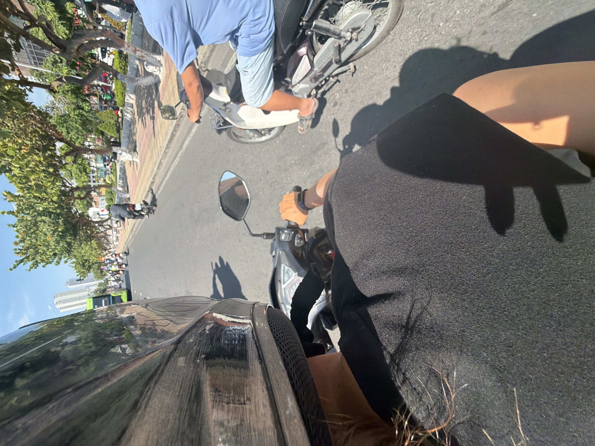 Captured from a first-person perspective on a moving motorcycle, the image portrays a street scene in Nha Trang Ward, Vietnam. Another motorcycle, driven by a person wearing a light blue shirt and light-colored shorts, is positioned ahead on the right side of the road. Buildings and trees line the street's left side, with a glimpse of a bus and tall buildings visible in the distance. The asphalt road is visible, and the overall lighting suggests a sunny day.
