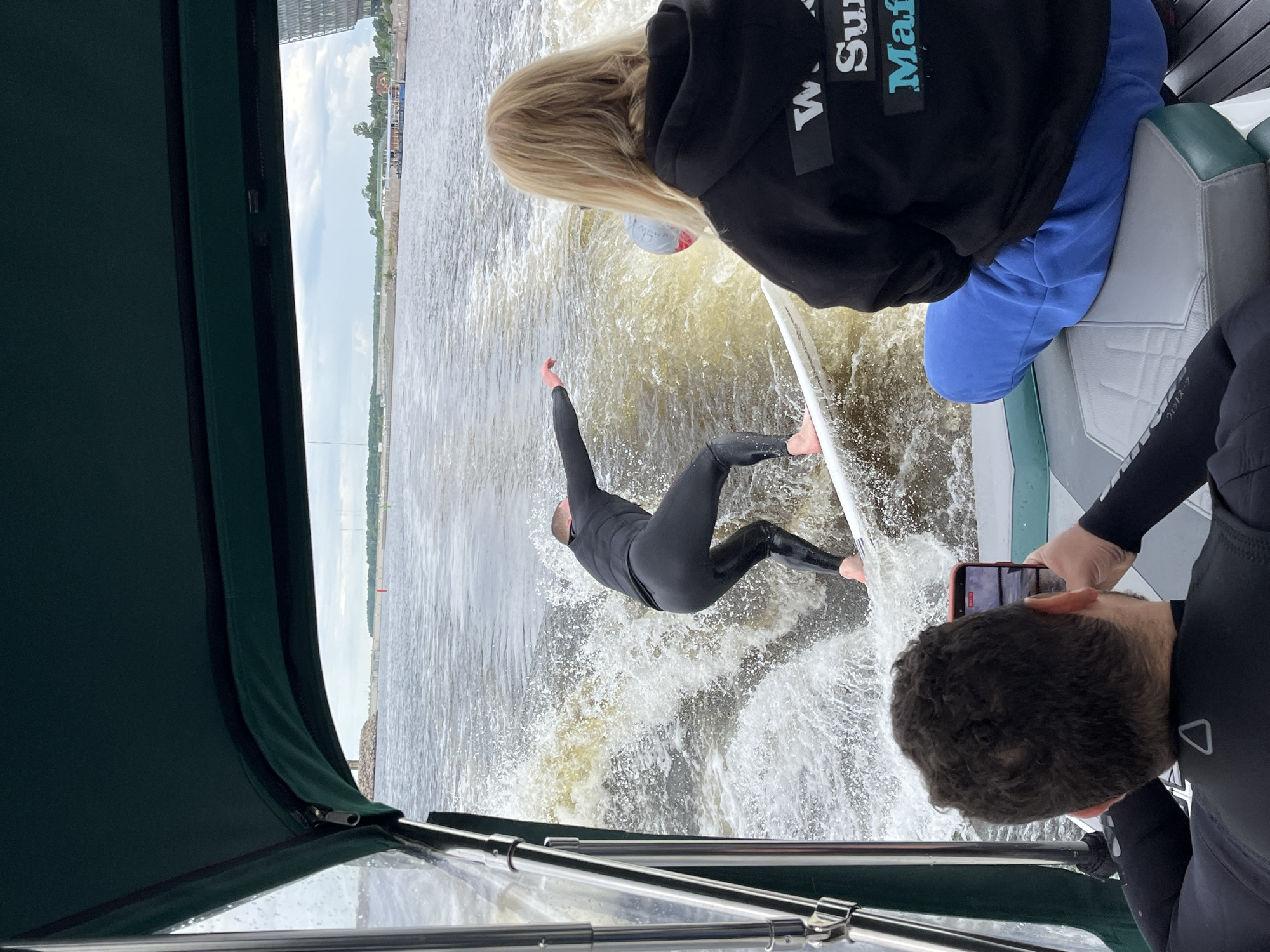 An over-the-shoulder shot shows three people on a boat as it speeds through the water, creating a wake. In the foreground, a person wearing a black wetsuit is seated, holding a smartphone to record. To the right, another person wearing a black hoodie and blue sweatpants is seated, looking towards the wake. In the center, a person in a black wetsuit is standing on a surfboard, being towed by the boat. The water is choppy and brown, and the sky is overcast. The boat appears to be on the Neva River, near the Petrogradsky District of Saint Petersburg, Russia, with the modern buildings of the Lakhta Center visible in the distance on the right horizon. The boat has a green canopy, and the seating appears to be upholstered in grey and teal.