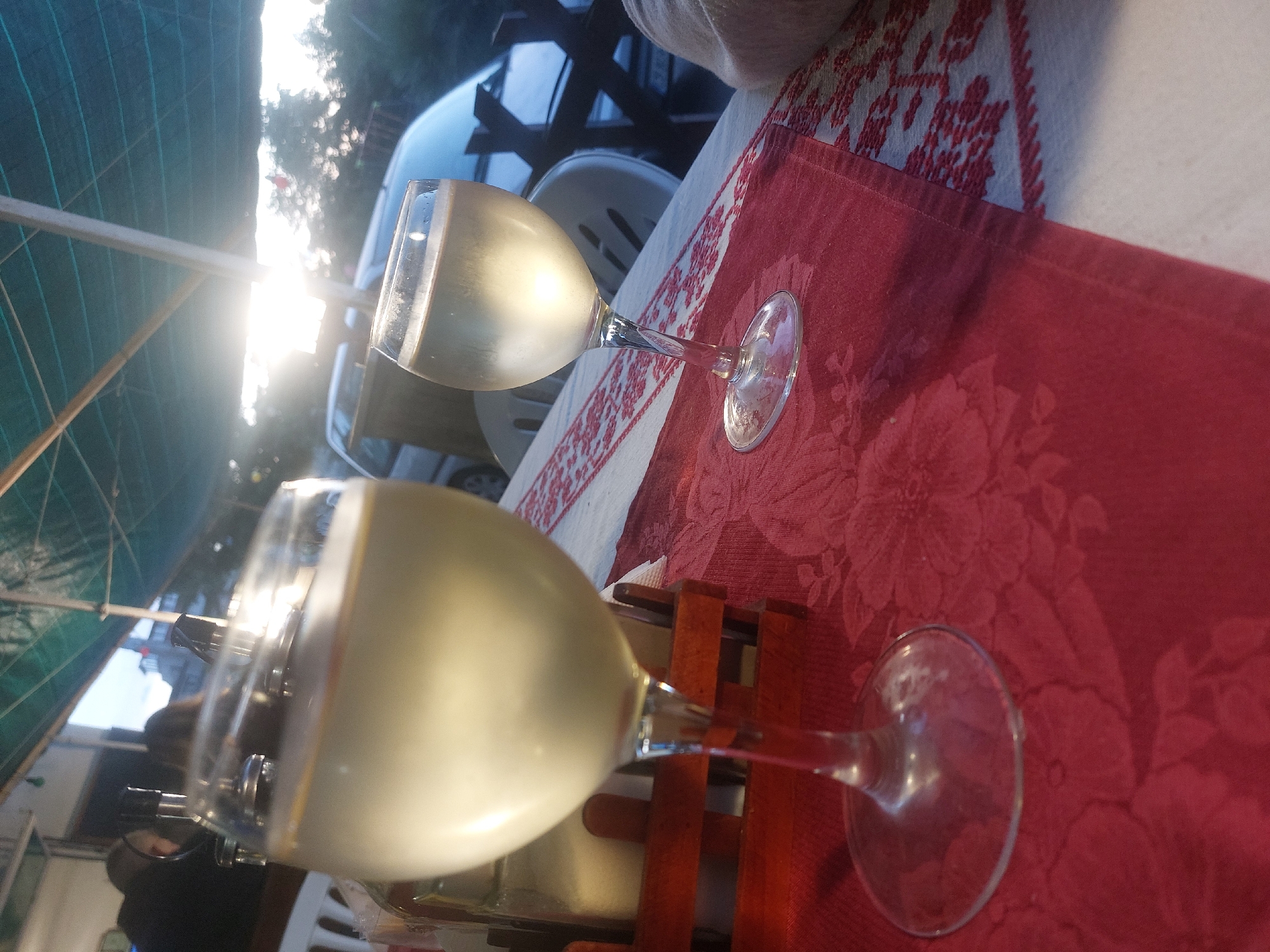 Here is a description of the image:
The image depicts two wine glasses filled with a light-colored liquid, presumably wine, on a table covered with a red, floral-patterned cloth. The glasses appear to be placed on a wooden wine rack. In the background, there are glimpses of the outside, including a vehicle and what appears to be a partially visible structure, possibly a tent or awning. The overall lighting suggests it's daytime, possibly during sunset. The location, specified as Tsarevo, Bulgaria, is not directly evident in the image but assumed as the context. There are no people visible in the immediate foreground of the image, though there is a hint of a person in the far background.