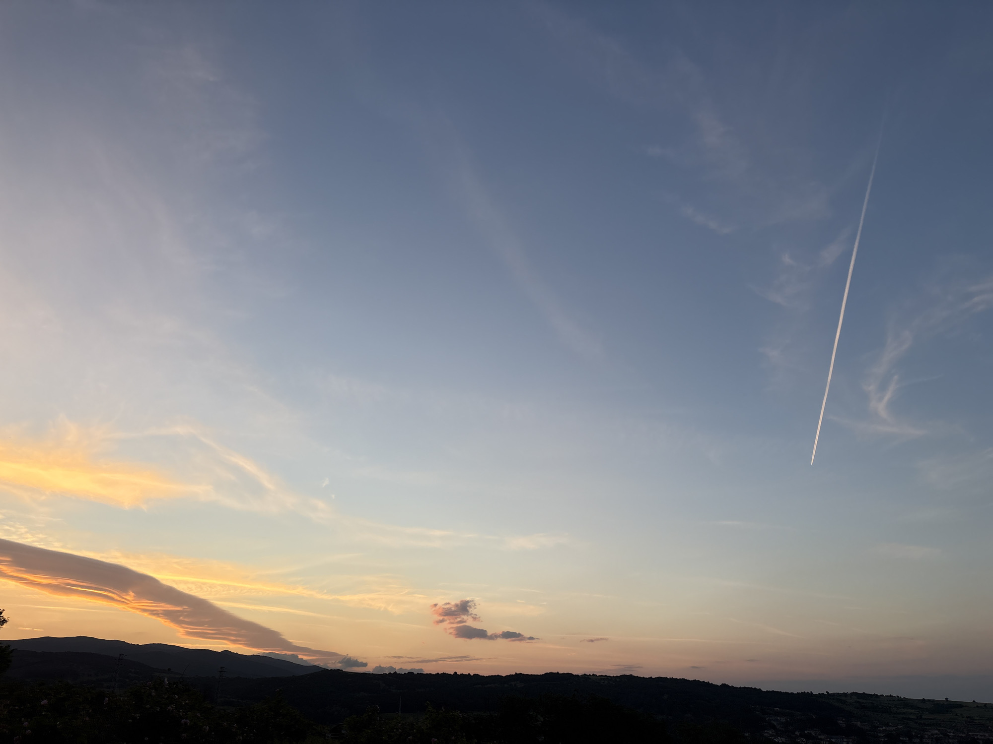 The image captures a landscape scene in Strelcha, Bulgaria, showcasing a tranquil sunset. The horizon line is dominated by a silhouetted range of hills and trees. The sky transitions from a light blue at the top to shades of orange and yellow near the horizon. Several cloud formations are present, including a long, finger-like cloud illuminated by the sunset and a small, pink-tinted cloud near the horizon. A jet contrail cuts vertically through the upper right portion of the sky. The lower part of the image is darker, likely due to shadow and vegetation, with hints of buildings possibly indicating residential areas.