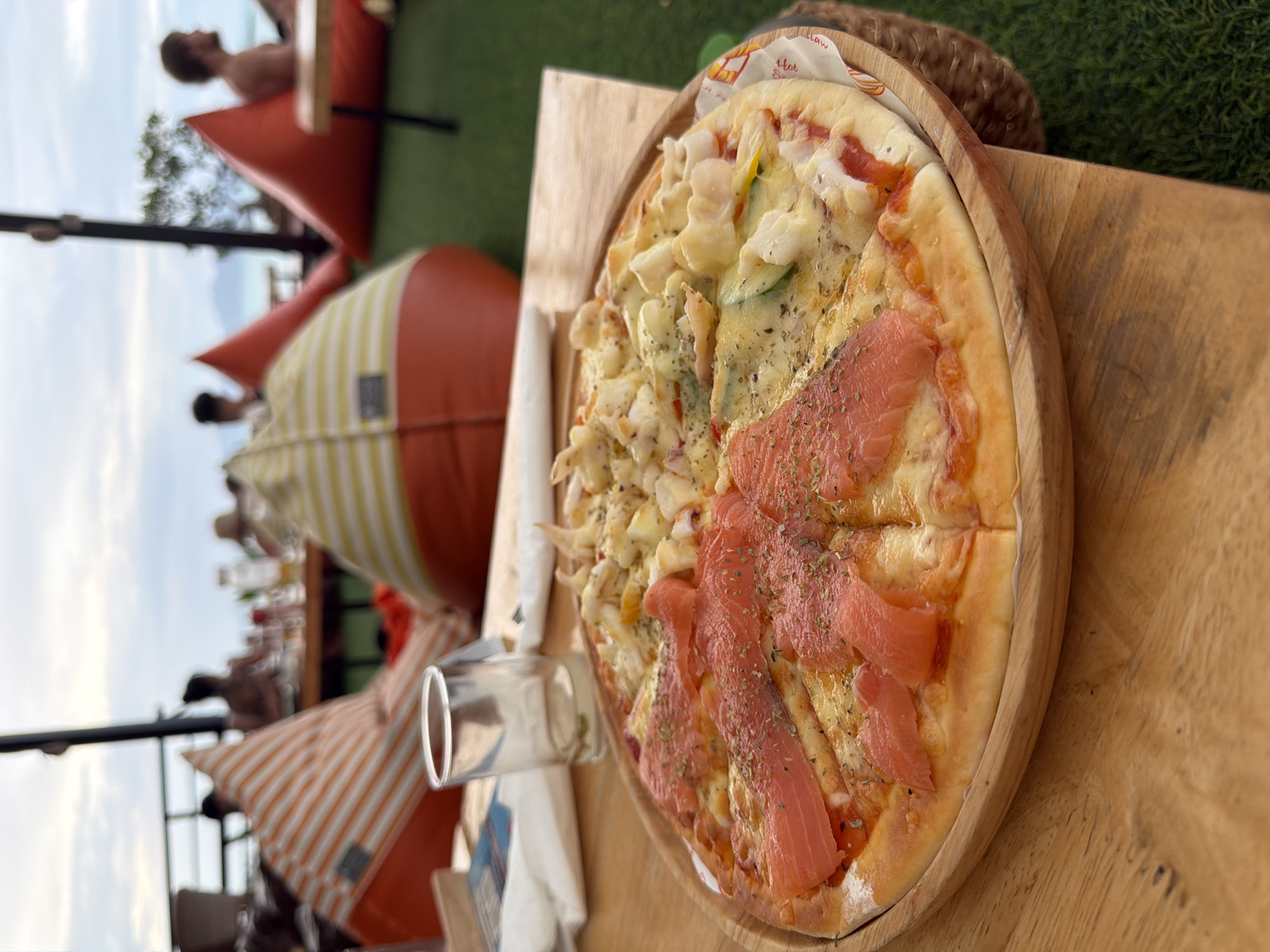 A high-angle, eye-level shot shows a close-up of a pizza with seafood and salmon toppings on a wooden plate, placed on a wooden table. The pizza is divided into four sections, with two sections covered in salmon and the other two with seafood, including shrimp and calamari, and possibly some vegetables such as zucchini. The crust appears to be thin and golden brown. To the left of the pizza, a clear glass with a light-colored liquid and some green specks is visible. In the background, out of focus, are people seated on beanbag chairs on a green lawn. The scene suggests a relaxed, outdoor dining setting, likely a restaurant or beach bar in Ko Samui District, Thailand, given the tropical attire of the patrons and the coastal vibe. The sky is overcast with a pale blue hue. The overall ambiance is casual and vacation-like.