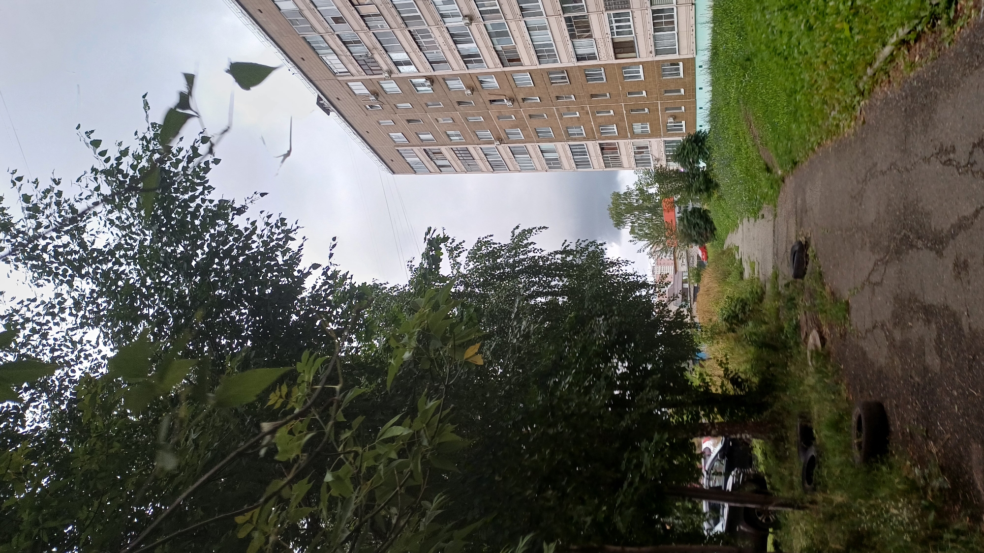 The image depicts a cloudy day scene in Izhevsk, Russia, featuring a long, multi-story apartment building with a beige facade and numerous windows. To the left of the building, a paved walkway runs alongside a grassy embankment, bordered by lush green trees with dark leaves. In the foreground, the walkway is lined with tires scattered on the grass. The background shows more trees and buildings, suggesting a residential area. The overall atmosphere is overcast and somewhat somber.