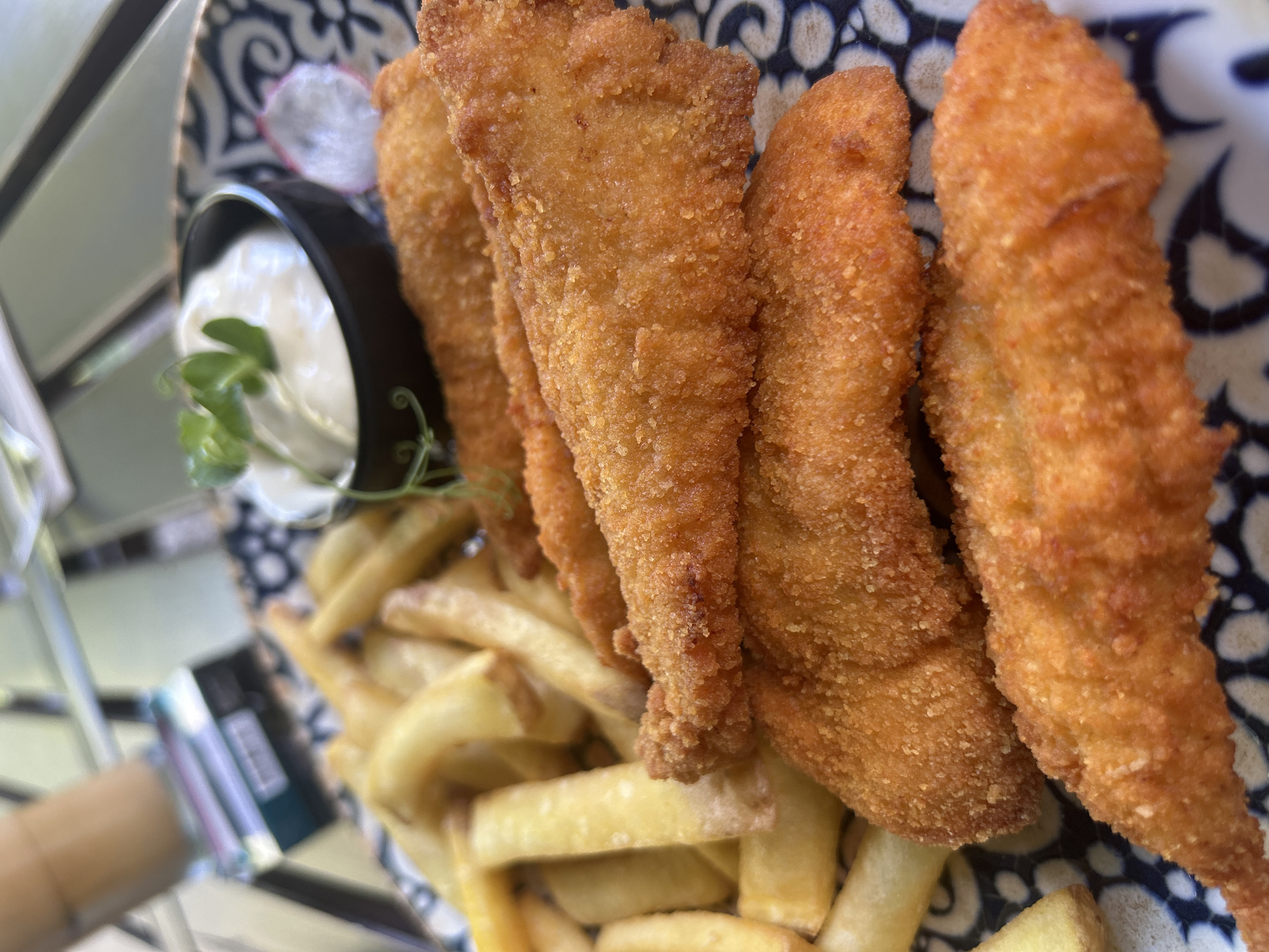 A plate of fried fish pieces and french fries is served with a small bowl of white sauce and a sprig of green garnish. The plate has a blue and white patterned rim and rests on a table outdoors. In the background, a blurred cigarette pack and a drink are visible. The scene appears to be at a restaurant in Varna, Bulgaria.