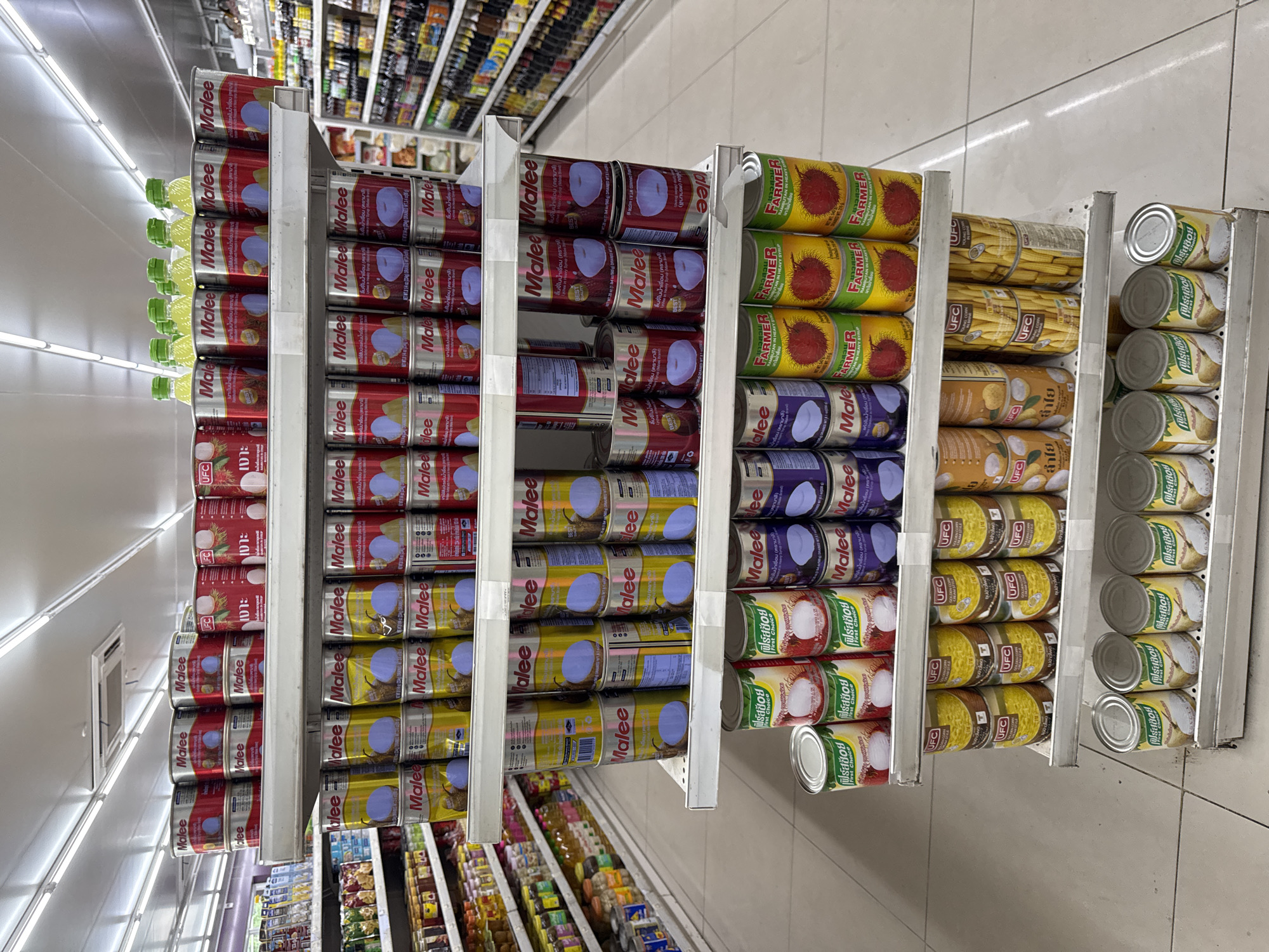 The image shows a grocery store shelf in what is likely Thong Sala, Thailand, stacked with canned goods. The brands "Malee" and "UFC" are prominently displayed. The cans include lychees, rambutan, corn, and longan fruits. The shelves are white metal, and the floor is a light gray tile. There are fluorescent lights overhead.