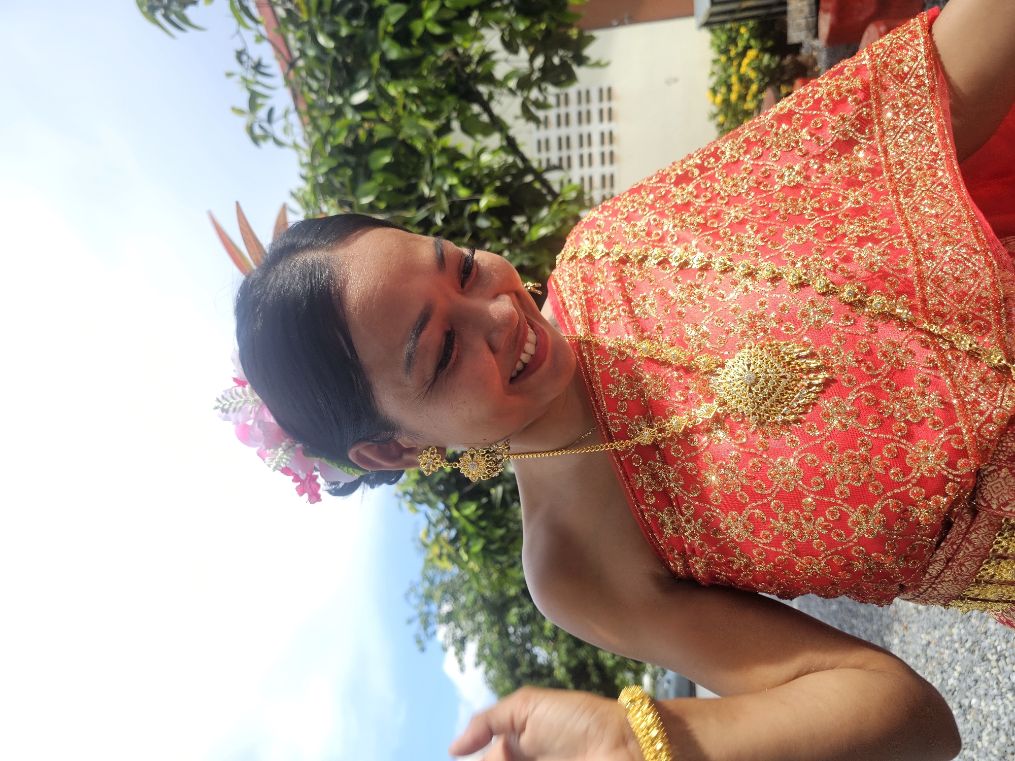 A medium shot shows a smiling Southeast Asian woman in traditional Thai attire, possibly for a ceremony or festival, outdoors under a bright, sunny sky. She is positioned in the foreground, her body angled slightly to the right, with her head tilted down and her gaze directed towards her left. Her dark hair is pulled back and adorned with a pink flower crown. She wears a vibrant red, one-shoulder garment heavily embellished with gold embroidery and sequins. Around her neck, a large, ornate gold pendant hangs from a chain, complemented by matching gold earrings and a thick gold bracelet on her left wrist. Her right arm is bent at the elbow, hand held up near her face, and her left arm is mostly out of frame. The background features lush green foliage, a building with horizontal vent details, and a hint of a yellow flowering plant. The ground appears to be covered with small, light-colored gravel. The overall impression is one of joy and cultural celebration in a tropical setting. The image was captured in Thep Krasatti, Thailand.