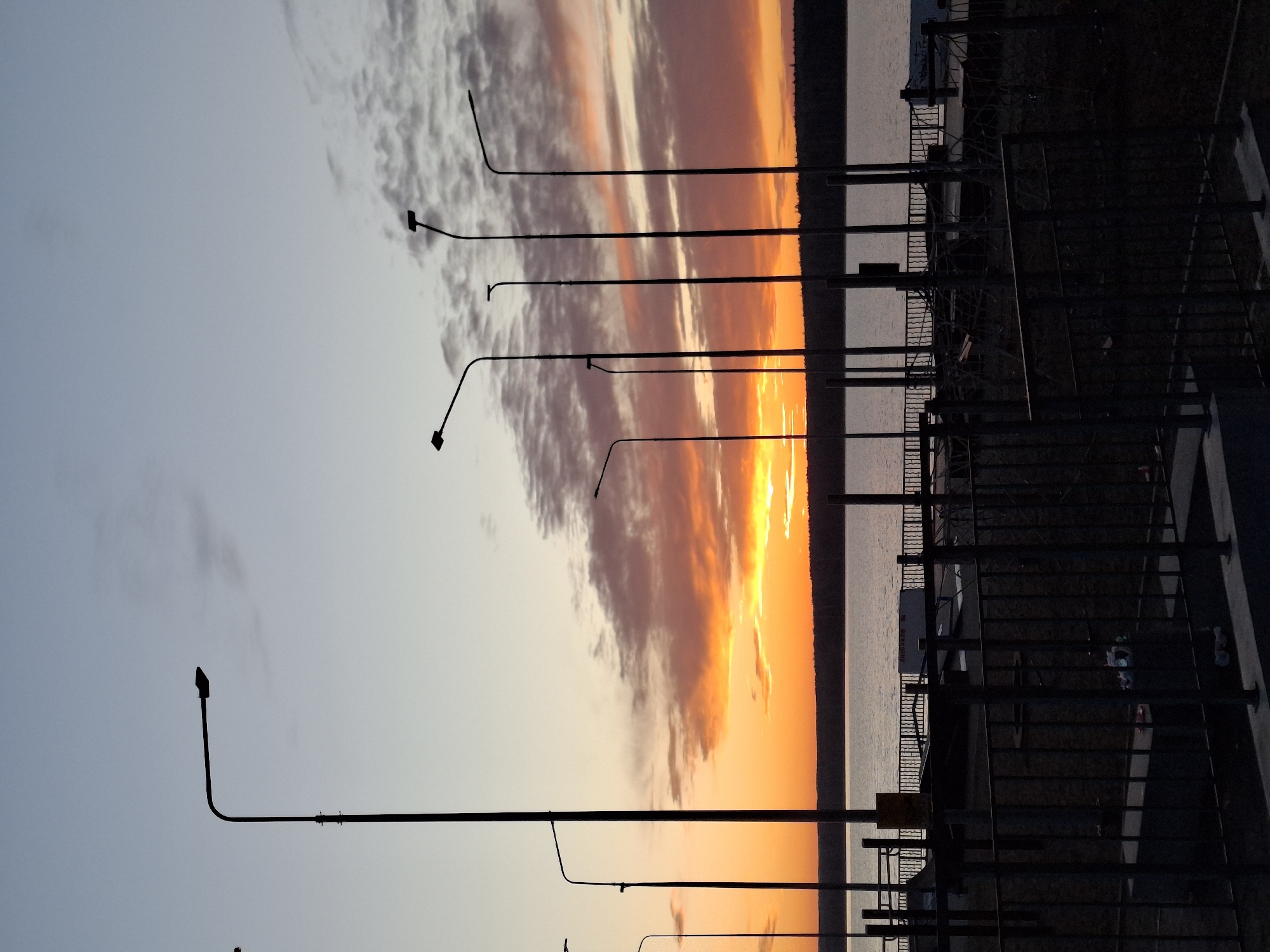 The image captures a sunset scene in Chaikovsky, Russia, featuring a waterfront area with lampposts and metal fencing. The sky is a gradient of colors, transitioning from light blue to orange and yellow where the sun is setting. Clouds are visible, adding texture to the sky. Silhouetted lampposts line the waterfront, extending into the distance. The metal fencing, also silhouetted, runs along the edge of what appears to be a walkway or promenade. The water's edge is visible, with some distant land or structures on the horizon. The image is taken at dusk, with the light primarily coming from the setting sun.