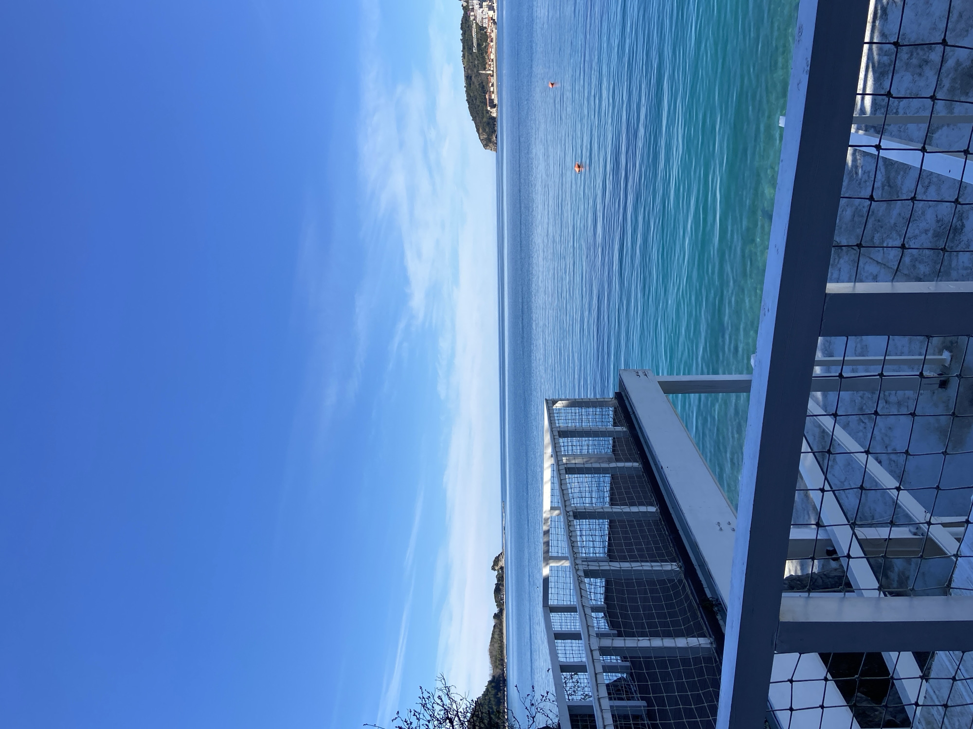 The image captures a serene coastal scene in Budva, Montenegro. The clear turquoise water of the Adriatic Sea stretches towards the horizon, punctuated by two orange buoys floating in the distance. A headland with buildings is visible on the horizon. A metal staircase with a mesh safety barrier dominates the right side of the frame. The sky is a bright blue with wispy clouds.