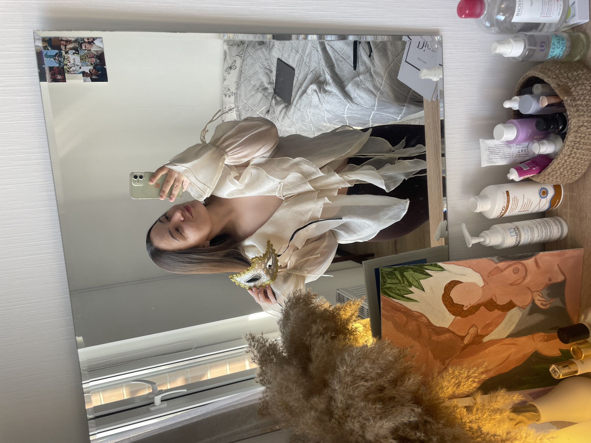 A young woman is taking a selfie in a mirror. She is wearing a white, flowing blouse and holding a white and gold mask. The background shows a bed with gray linens and a tablet on it. To the right of the mirror, there is a collection of cosmetic products, including bottles, tubes, and a basket containing smaller items. A painting with a nude figure is also visible. A vase with dried pampas grass stands in front of the painting. The location may be an apartment in Balashikha, Russia, based on the presence of Russian-labeled cosmetic products, but that cannot be determined with certainty from the image.