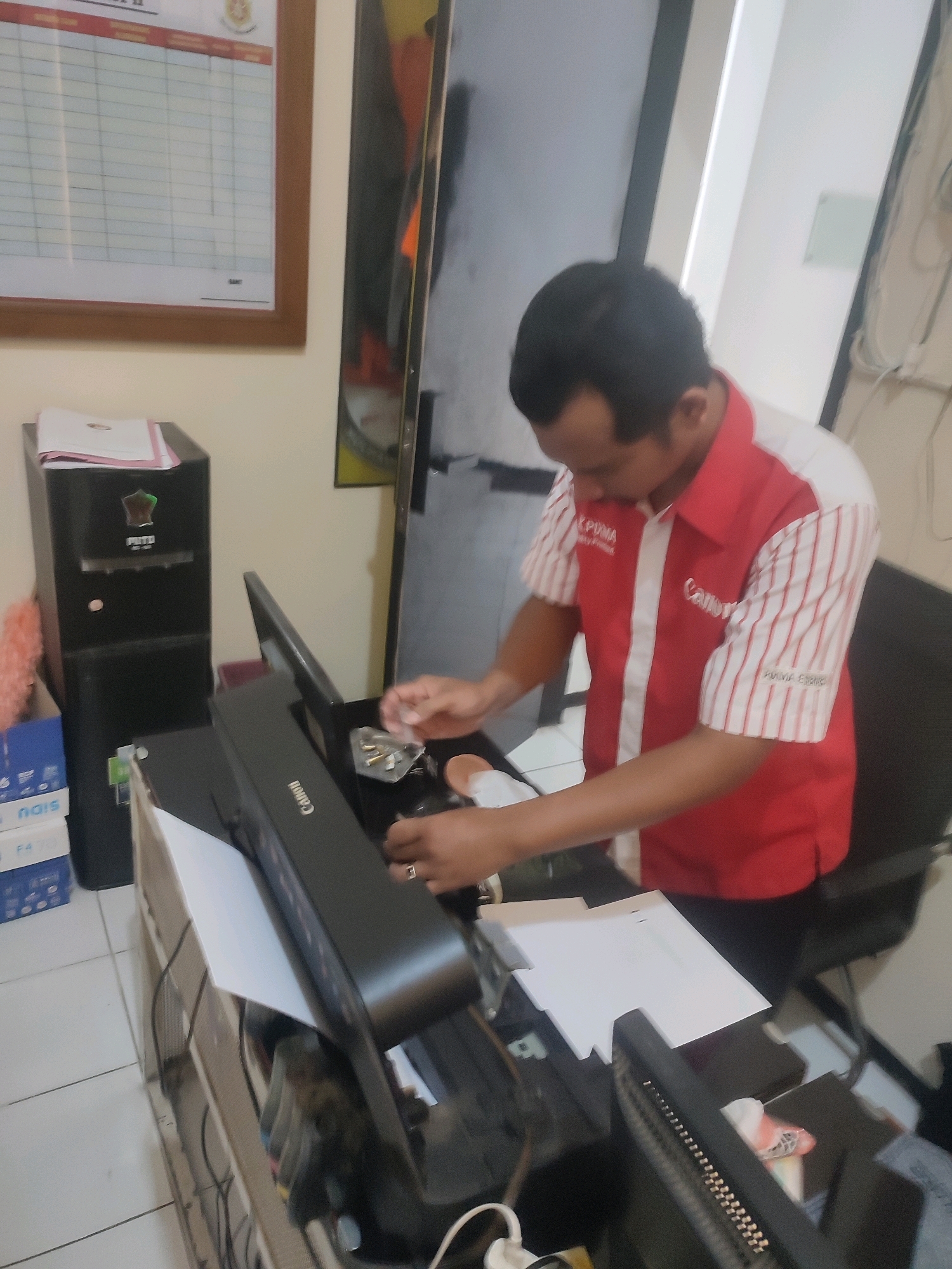 A man in a red and white striped shirt, likely a uniform with "Canon" branding, is working on a computer printer. The man has short, dark hair and is looking down at the printer. He appears to be holding a small tool or part in his right hand, as if making a repair or adjustment. The printer is black and has "Canon" written on it. To the left of the printer is a black computer tower with a green sticker. In the background, there is a whiteboard or bulletin board with a grid-like structure and a wooden frame. The environment appears to be an office or workshop setting, with office supplies and equipment visible. The floor is tiled. The scene is dimly lit, with some natural light coming from the right side of the image.