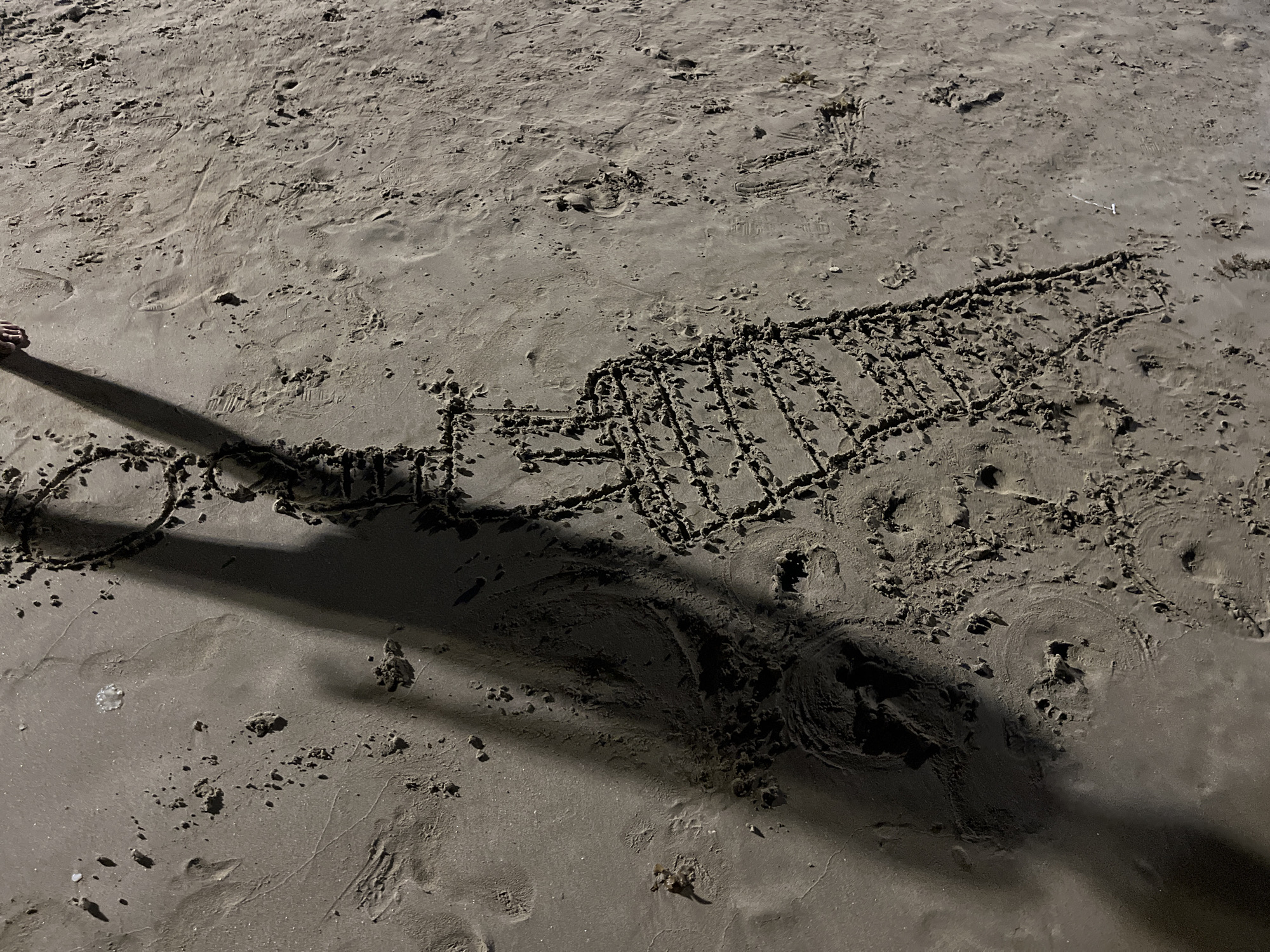 The image shows a drawing of a large truck, possibly a semi-truck, in the sand. A shadow of a person's leg and foot is visible on the left side of the image, suggesting the person is standing nearby. The scene is on a sandy beach.