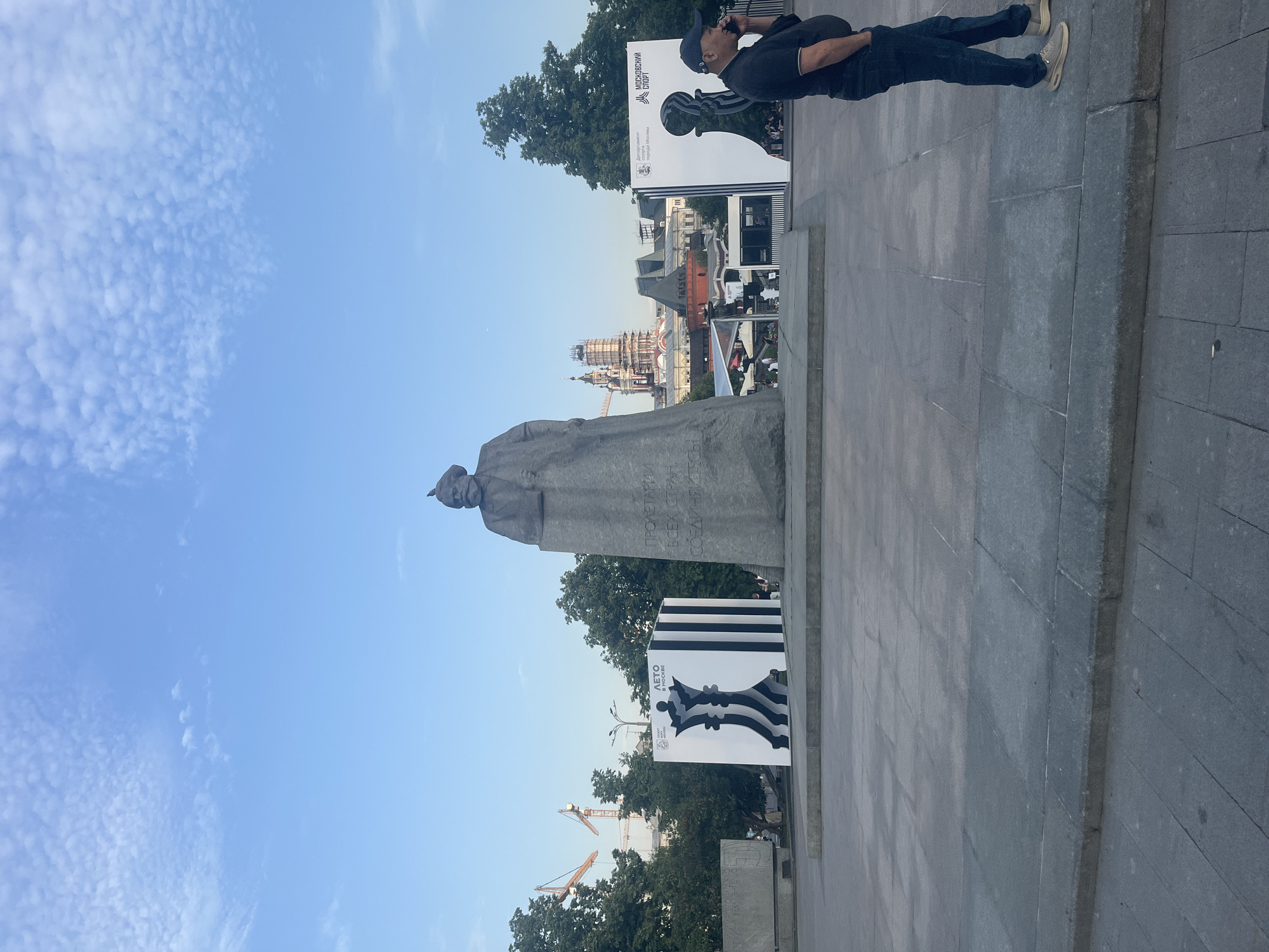 This is a wide, eye-level, outdoor, daytime photograph taken in Moscow, Russia. The image features a large stone monument of Karl Marx in the foreground, with Cyrillic text inscribed on its base. To the left and right of the monument are large, stylized black and white chess pieces, one depicting a queen with the word "ЛЕТО" (Summer) and another with the Moscow coat of arms and the words "МОСКОВСКИЙ СПОРТ" (Moscow Sport).
A man in a dark polo shirt, blue jeans, and a baseball cap is standing on the right side of the image, holding a phone to his ear. Behind the monument, a portion of Moscow's skyline is visible, including historical buildings with ornate red and white architecture and church domes, as well as construction cranes. The sky is blue with scattered, wispy clouds. The ground is paved with large, gray stone slabs. The overall scene appears to be a public square or park.