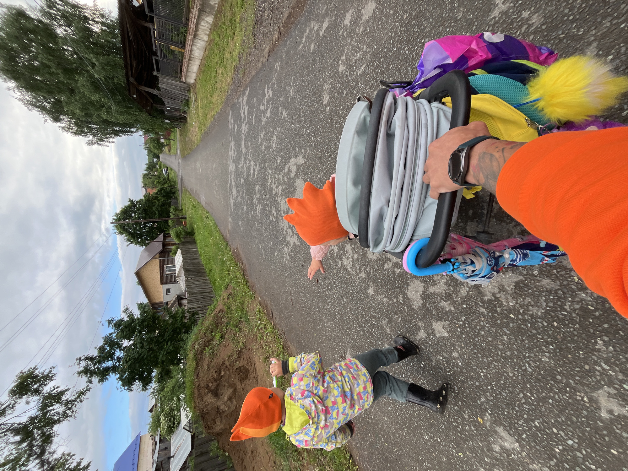 The image, apparently a selfie taken by a person in an orange long-sleeved shirt, shows a child in a colorful jacket, dark trousers, and black boots, walking on a paved road. The child wears a pointed orange hat. Beside the child, the person is pushing a stroller or buggy laden with folded fabric and miscellaneous colorful items. Another child, visible only by their hand and part of their head, also wearing an orange hat, is near the stroller. The person's arm and hand, with a watch visible, are in the right foreground. The road stretches ahead, lined with grass and vegetation on either side. In the background, there are houses and trees. The location is specified as Chaikovsky, Russia. The sky is overcast.
