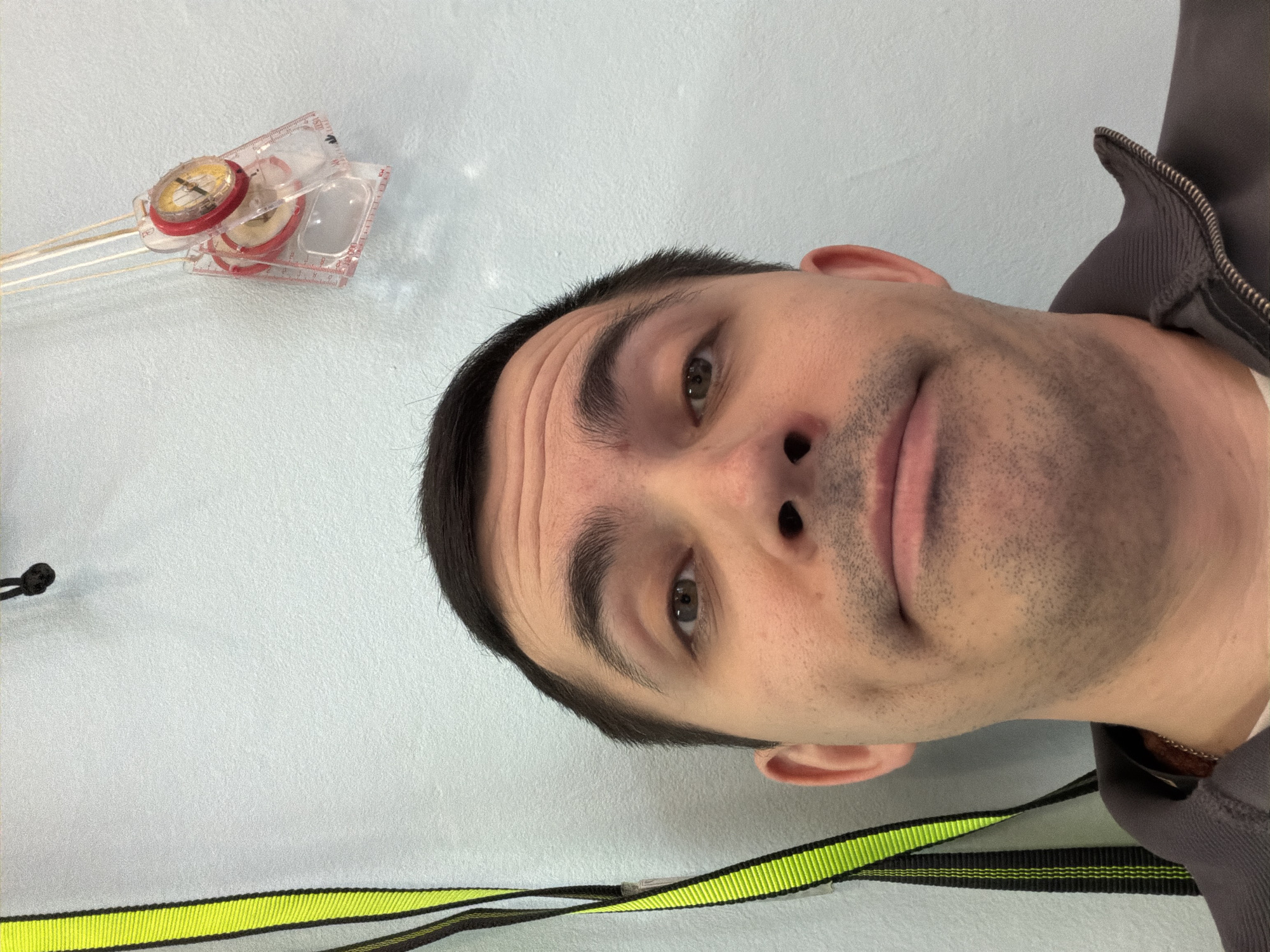 The image captures a close-up, somewhat tilted portrait of a man, seemingly a selfie, against a light-colored, possibly light blue or gray, wall. The man, with short dark hair and visible stubble, looks towards the camera with a neutral to slightly curious expression, his brows subtly furrowed, creating horizontal lines on his forehead. His brown eyes are open, and his mouth is closed, with his lips appearing slightly parted. He is wearing a gray zippered garment, possibly a jacket or hoodie, visible on his right shoulder and neck.
In the upper left corner of the frame, positioned on the wall, are several small objects. These include a clear plastic ruler with numerical markings (e.g., "1", "2") and two or three small, round compasses with red rims and yellowish dials, attached to thin, white sticks. They appear to be educational or craft items.
Along the bottom edge of the image, a black strap with prominent neon yellow horizontal stripes runs horizontally. This strap might be part of a lanyard, a harness, or a bag strap. The lighting suggests an indoor setting, and no specific time of day is discernible. The man's gaze and the angle of the shot give a candid feel to the portrait. There are no explicit location cues in the image itself, though the provided context indicates the location is Izhevsk, Russia.