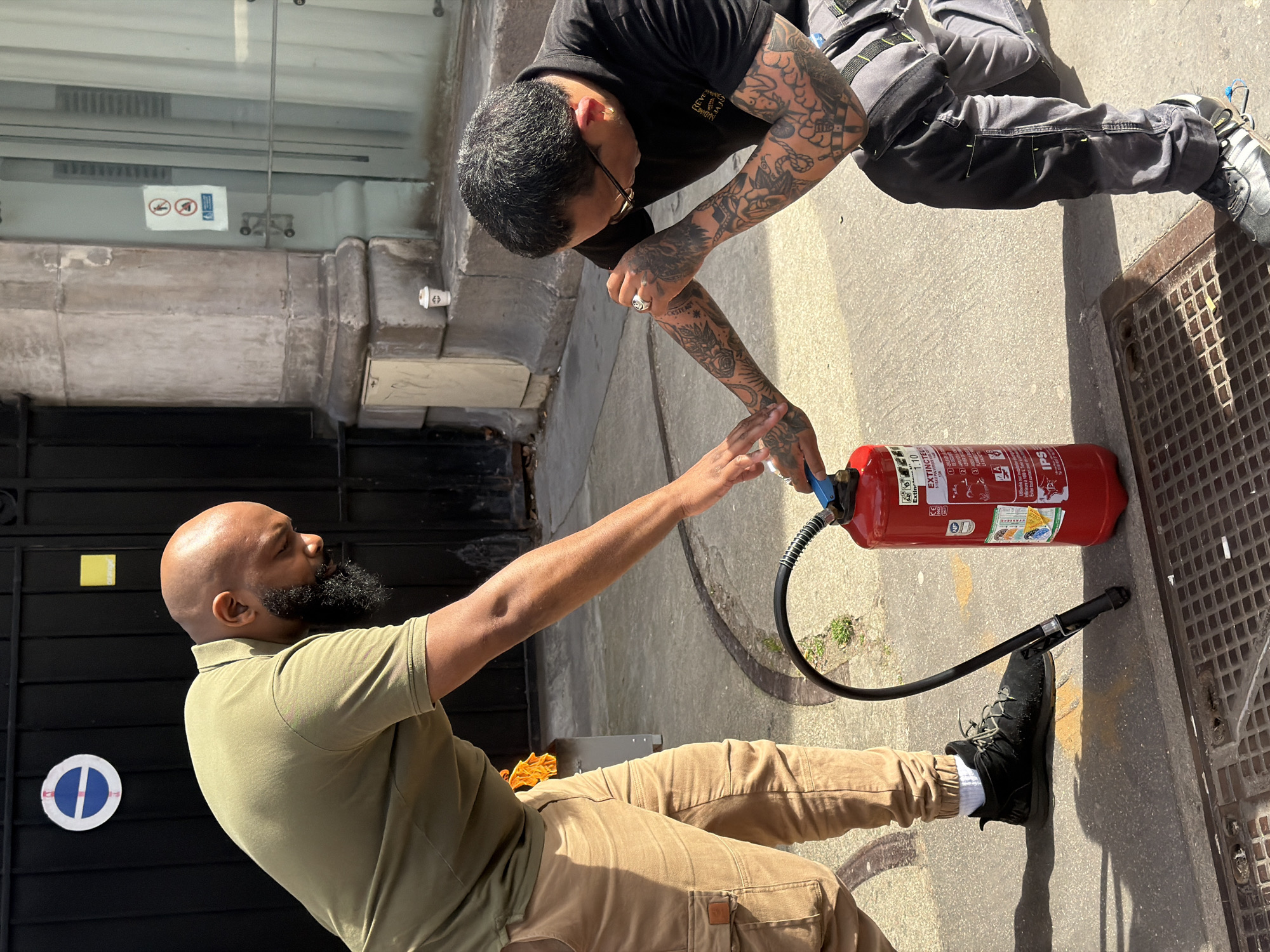 Two men are interacting with a red fire extinguisher on a sidewalk in what appears to be an urban environment. The man on the left is kneeling, wearing a green shirt and khaki pants, and has a bald head and a dark beard. He is extending his arm towards the extinguisher. The man on the right is leaning over the extinguisher, wearing a black t-shirt, gray work pants with black knee pads, and is heavily tattooed on his arms. The fire extinguisher is lying horizontally on the sidewalk next to a drain cover. A black hose is connected to the extinguisher. The background includes a building with a dark door and stone architectural details. Reflected signage, which appears to prohibit parking and has a no-smoking emblem, is seen on the building. Based on the architectural style and signage, the location is potentially in the city of Paris, France.