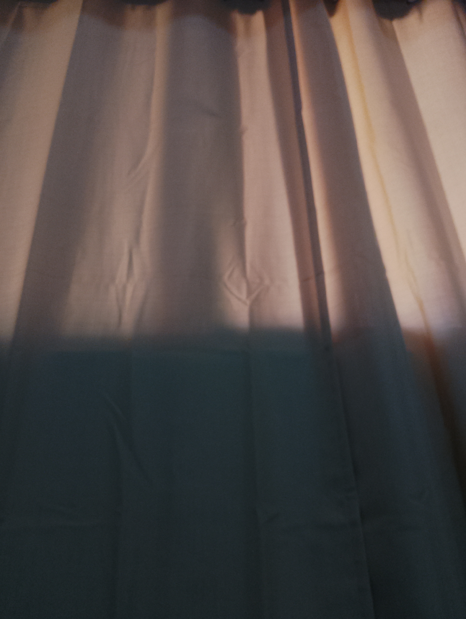 The image shows a close-up view of off-white curtains. The top portion of the curtains is illuminated by sunlight or artificial light, while the lower portion is shadowed. The lighting creates distinct vertical lines and folds in the fabric. There is no indication of location or any identifiable person, subject, object, or interaction in the environment. It cannot be said that this picture was taken in Pampatar, Venezuela.