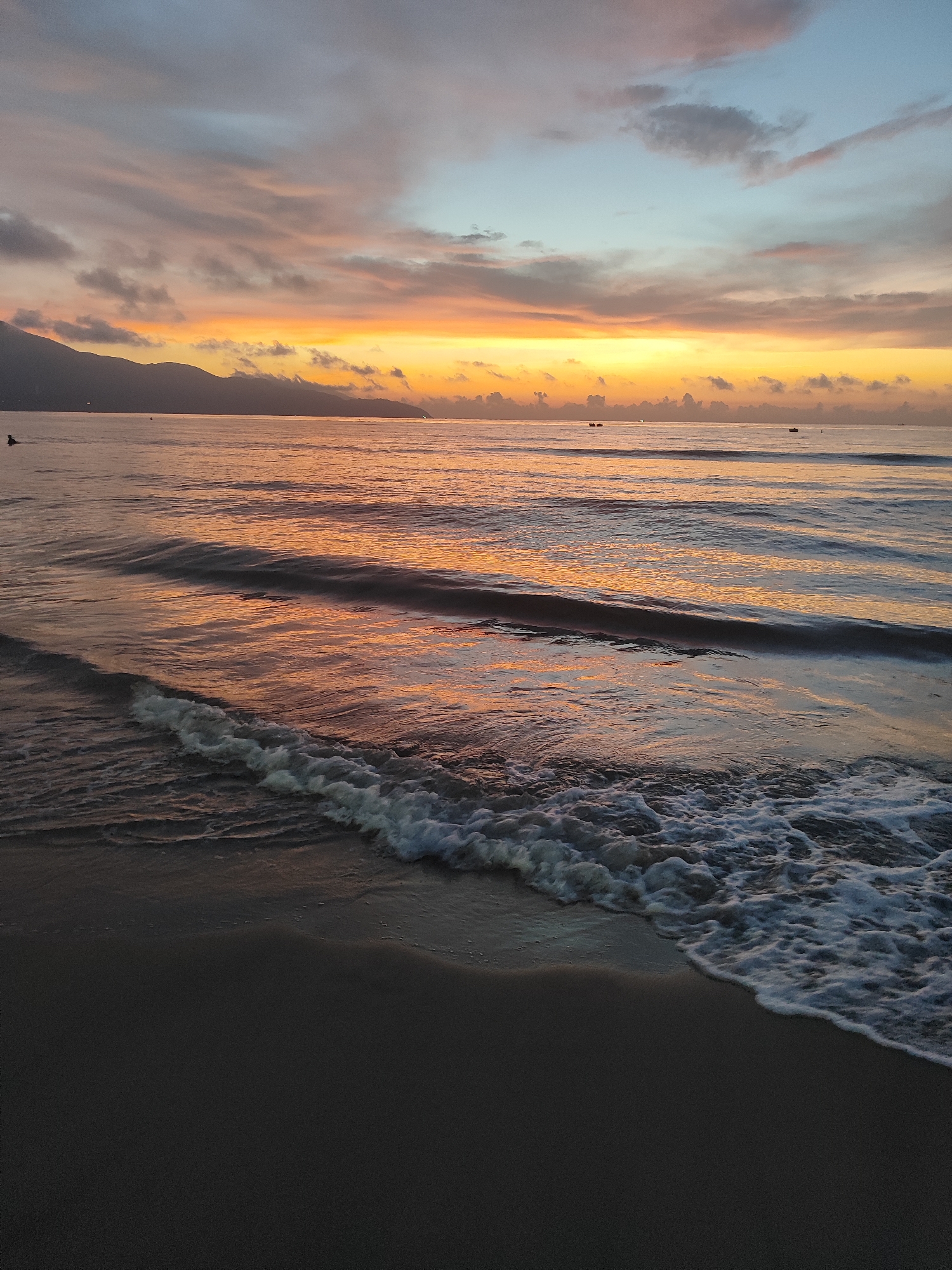 The image depicts a beach scene at sunrise or sunset, likely in Đà Nẵng, Vietnam, due to the presence of mountains along the horizon. The sky is a mix of colors, with oranges and yellows closest to the horizon transitioning to pinks, blues, and grays higher up. Clouds are scattered across the sky. The sea reflects the colors of the sky, with a predominant orange hue. Waves are breaking on the shore in the foreground, with white foam visible. The beach sand in the foreground is dark. A few boats are discernible on the horizon. A person can be seen in the water on the left.