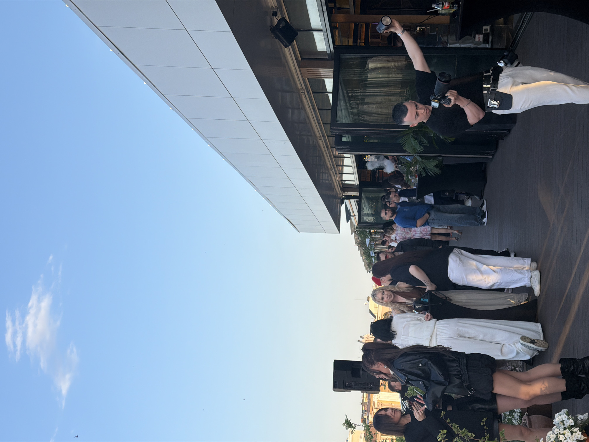 The image shows an outdoor event with a gathering of people in Kazan, Russia. A modern building with a reflective facade stands to the left, contrasting with the blue sky above. A photographer in white pants and a black shirt is capturing the scene. Below the building's edge, a crowd of people is assembled. Some are dressed in white outfits, others in casual wear such as jeans and blue shirts. A black speaker is visible near the front, and white flowers decorate the area. The overall atmosphere appears to be a public event with photography being a focal point.
