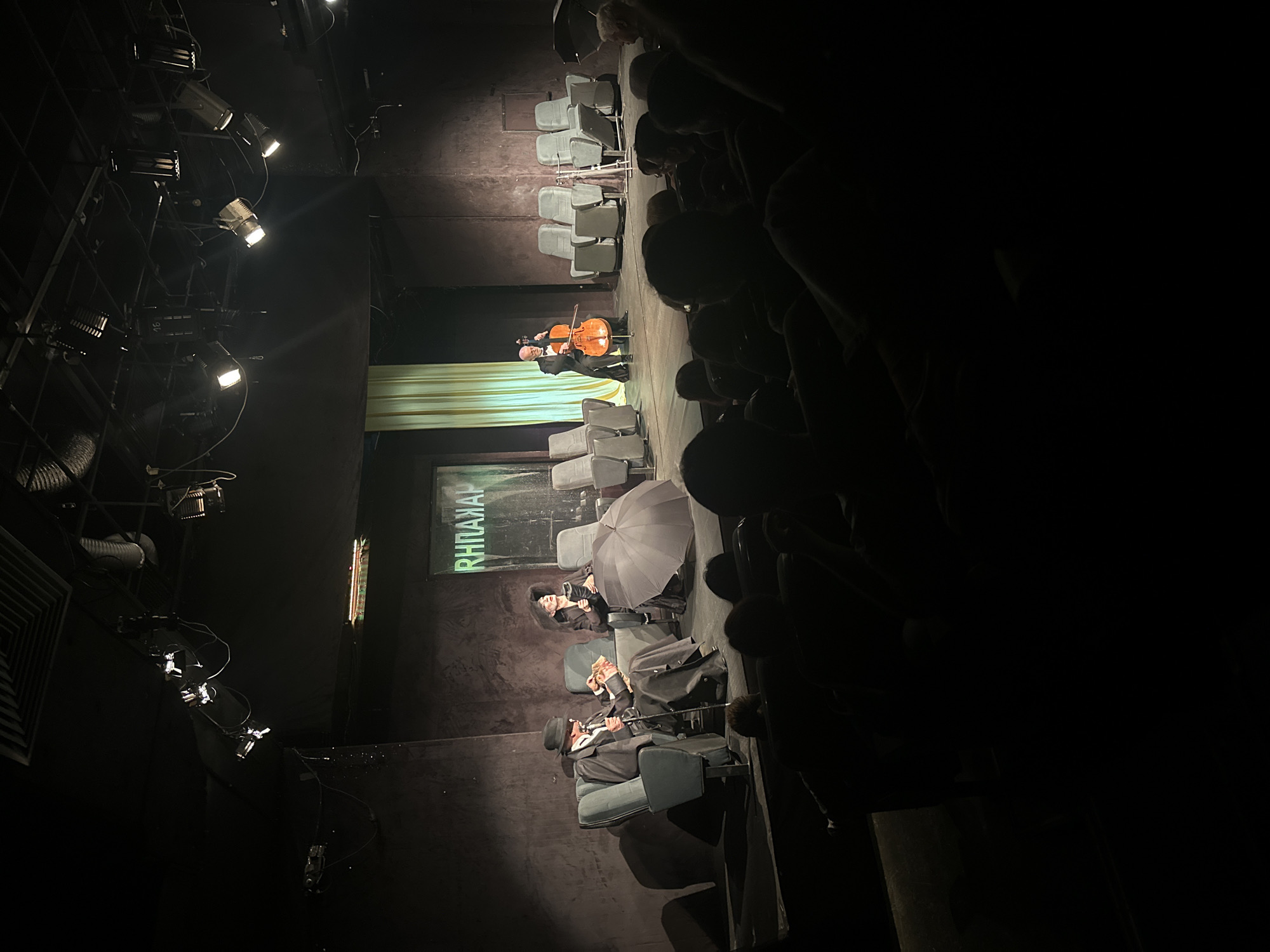 The image captures a scene from a theater performance, possibly in Sofia, Bulgaria, based on the Cyrillic text visible on the set piece. The foreground shows the audience sitting in a darkened theater, with figures silhouetted against the brighter stage. The stage features a performer playing a cello, illuminated by stage lights. Seated in the audience are two individuals with a distinct appearance, including a person holding an umbrella and another with a hat and cane. The set includes a green curtain and a sign with text in Cyrillic script. Additional stage lighting equipment is visible on the side of the stage. The overall atmosphere is dark and theatrical.