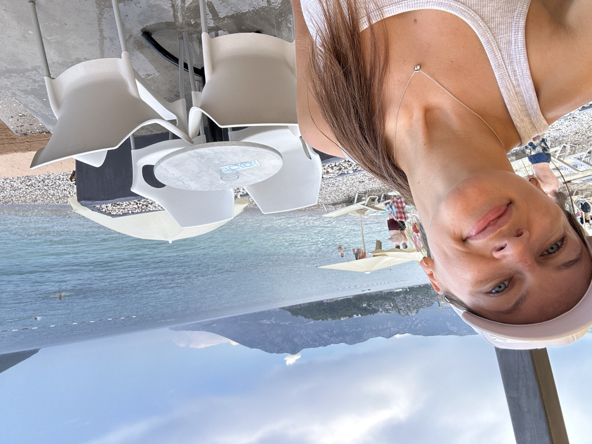 A woman in a white tank top and a white baseball cap takes a selfie in front of a beach. She has light blue eyes and brown hair pulled back. In the background, the sea meets a mountainous coastline. White chairs and a table with an ashtray are visible on a wet patio. Some beachgoers are in the water, and others are relaxing on lounge chairs under umbrellas. The location is identified as Budva, Montenegro.