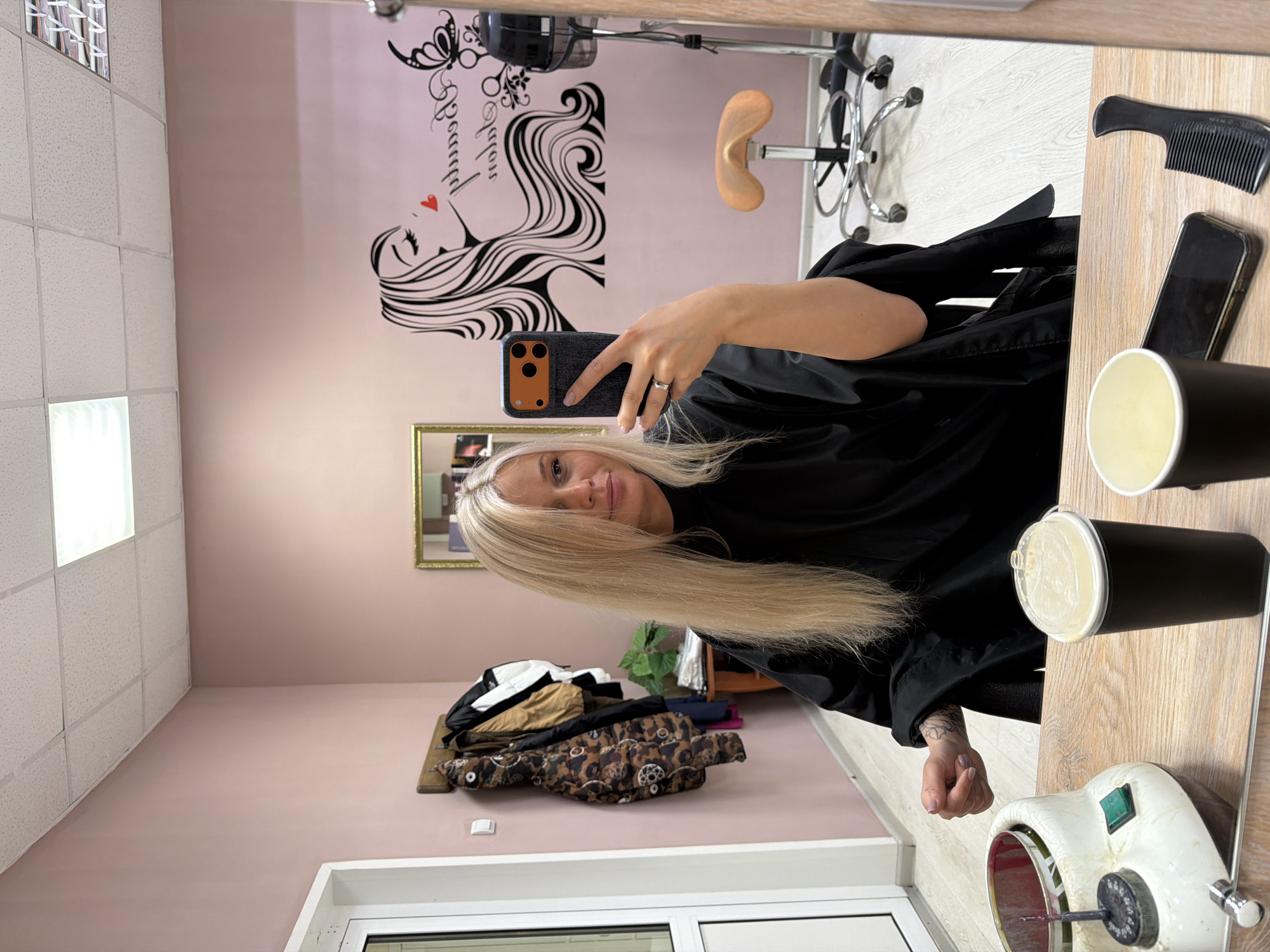 A woman with blonde hair, wearing a black salon cape, is taking a selfie in a hair salon. She is holding a smartphone with a black case and orange accents in her right hand. Her reflection is visible in a mirror in the background. The salon has light pink walls with a decorative illustration of a woman's profile with flowing hair on one wall. A salon chair with a brown seat is also visible in the background. On a counter in the foreground, there are salon tools including a black comb, a black smartphone, black containers, and a white cosmetic device with metal components. Clothes are piled on a surface near a window in the lower part of the image. The location is Sarapul, Russia, based on the location information.