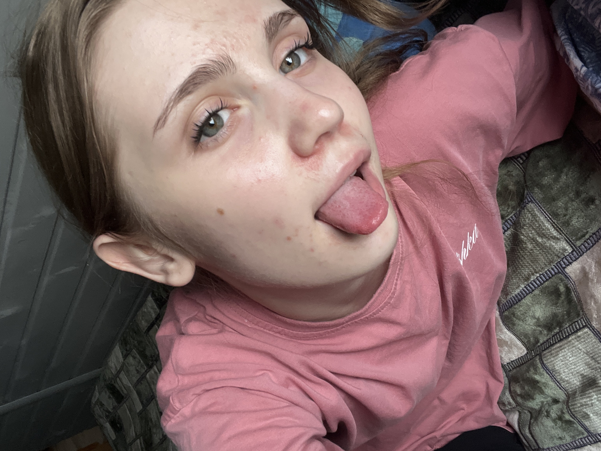 The media file is a close-up, high-angle selfie of a young person with light skin, green eyes, and light brown hair. They are wearing a pink long-sleeved shirt with a white embroidered logo on the left side of the chest, which appears to say "Vikka". The person is sticking their tongue out towards the camera, with their mouth slightly open. Their skin shows some redness and blemishes, particularly on the cheeks and forehead. Their eyelashes are dark and prominent.
The background is composed of a plaid blanket with green and dark blue squares and a dark grey ribbed surface on the left side. The lighting suggests an indoor setting, possibly in the daytime due to the natural light visible on the person's face. The overall mood appears playful and casual. The location is identified as Ashkadar, Russia. No specific activity or event is clearly depicted beyond the selfie being taken.