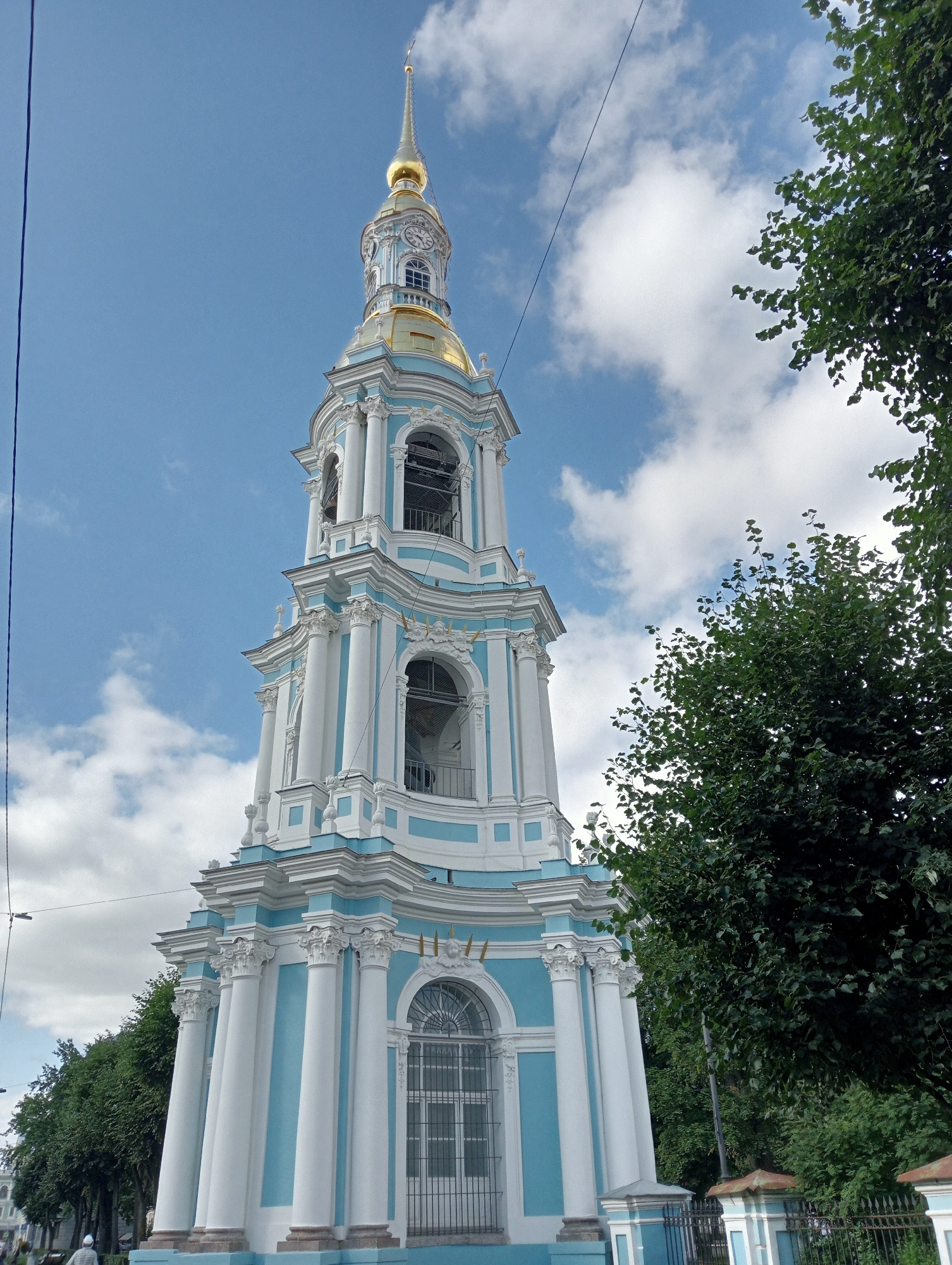 A tall, ornate bell tower with a gold spire stands against a blue sky with scattered white clouds. The tower is painted white and light blue, with white columns and decorative elements. Trees with dark green leaves flank the right side of the tower. In the lower left corner, a person is visible walking away from the camera, and a line of trees is visible along a street in the background. This is the bell tower of St. Nicholas Naval Cathedral in Saint Petersburg, Russia.