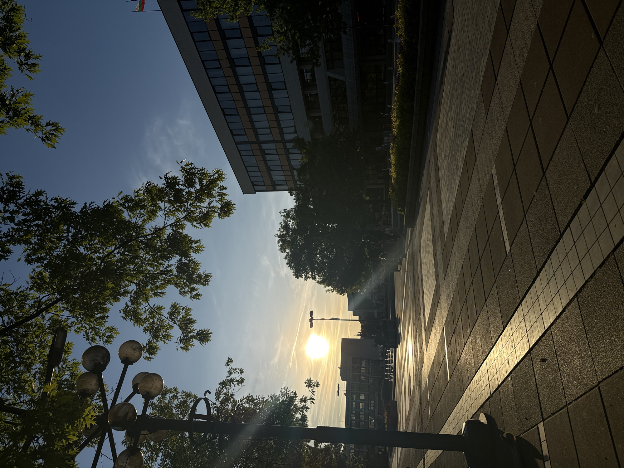The image shows a street scene in Blagoevgrad, Bulgaria, at what appears to be sunset or sunrise. The sun is visible in the distance, between buildings and trees, casting a glare. To the left of the frame is a tall lamp post with multiple spherical lights. Trees with green leaves are visible in the bottom left and top left corners. A multi-story building with a glass facade is in the upper part of the image. A flag can be seen flying on the building. A paved sidewalk extends from the bottom right towards the center. The lighting suggests a clear sky, with the sun being the dominant light source. No people are clearly visible in the scene.