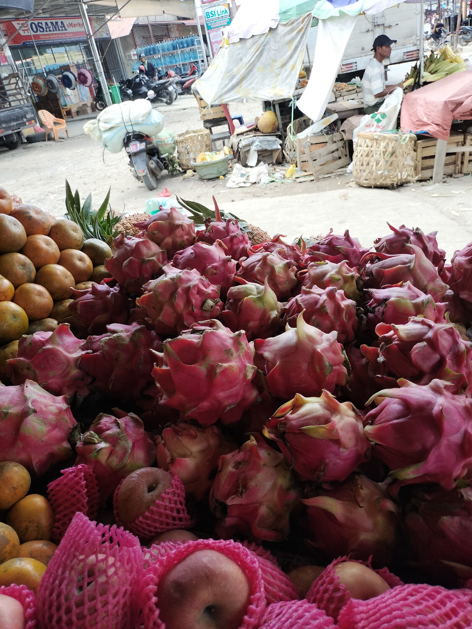 This is a photograph taken at a market in Kotapantonlabu, Indonesia. In the foreground, a large display of dragon fruits is prominently featured, their vibrant pink and green exteriors contrasting with the surrounding items. To the left of the dragon fruits, a mound of oranges is visible, some wrapped in pink netting. Behind the fruits, a collection of leafy green vegetables, possibly pineapples, is also present. The background reveals a bustling market environment. A man wearing a hat and striped shirt is seated behind a stall, attending to produce. Several motorcycles are parked nearby, one with a large load of what appears to be sacks tied to its back. Market stalls with awnings and various goods, including bottled water and other food items, are scattered throughout the scene. The ground is unpaved, typical of an outdoor market.
