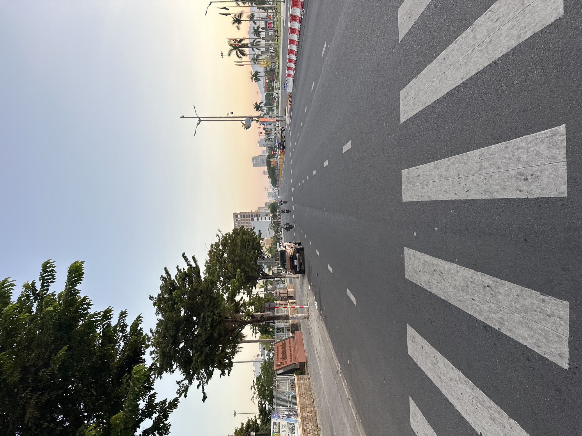 This is a wide-angle, eye-level shot of a street in Da Nang, Vietnam, possibly during dawn or dusk due to the warm, soft lighting of the sky. The foreground is dominated by a zebra crossing on a wide, multi-lane asphalt road. To the left of the road is a sidewalk with a low brick wall and trees. Beyond the wall, there is a large red sign with Vietnamese writing, a fence, and some posters. Several motorbikes and a car are visible on the road, moving away from the viewer. The background features a cityscape with tall buildings and palm trees, with mountains visible in the distance. Lampposts line the street, and a row of red and white traffic barriers is on the right side of the road. The sky transitions from a pale blue at the top to a soft orange and yellow near the horizon.
