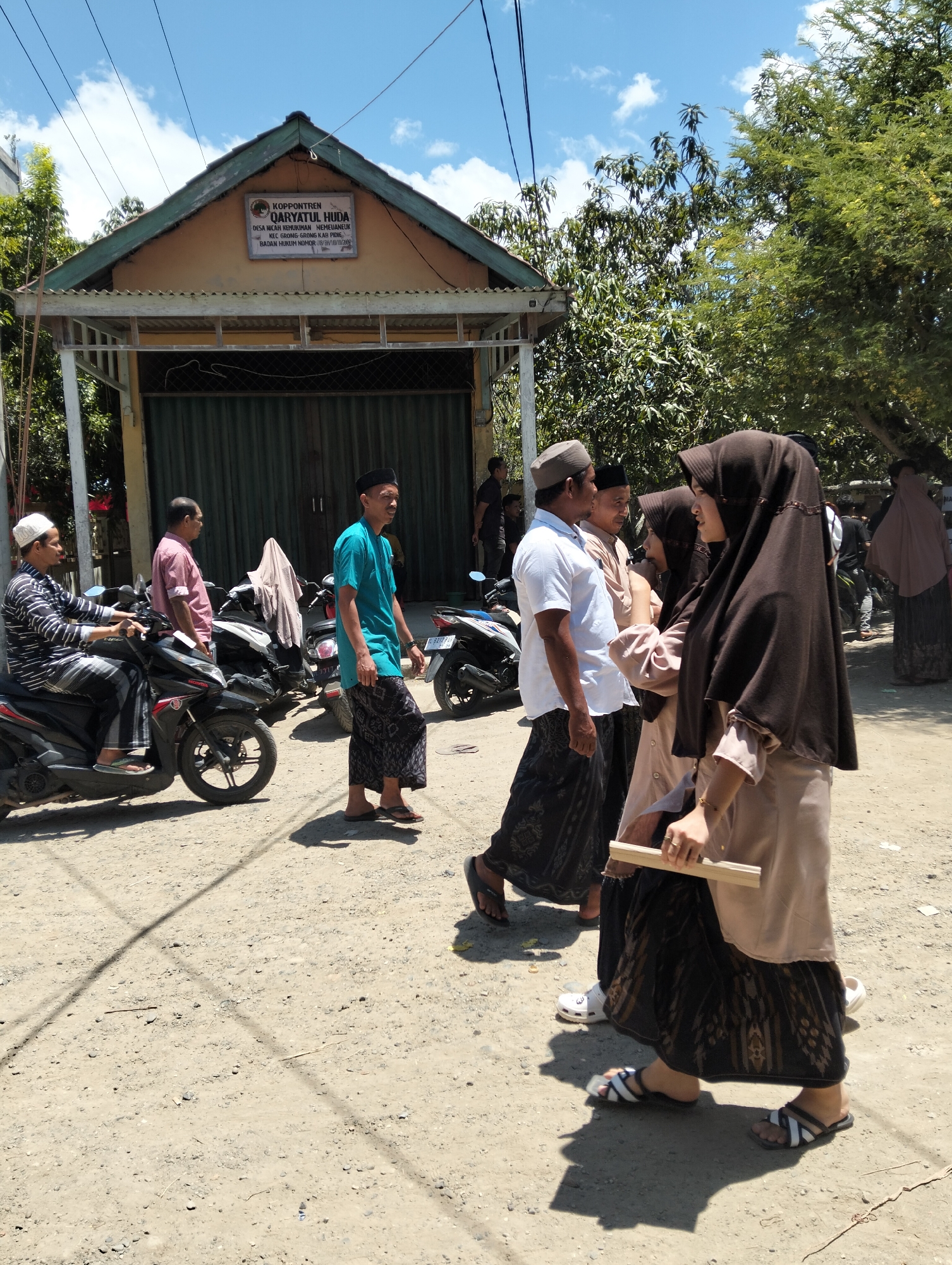 In the city of Teungoh, Indonesia, a group of people are gathered outdoors in front of a building. The building, likely a religious institution given the sign that reads "Koppontren Qaryatul Huda," has a yellow facade and a green roof. A large green metal gate covers the entrance.
Several men are present, some wearing traditional Islamic attire such as peci hats and sarongs. One man in a blue shirt and patterned sarong walks past the building, while another in a white shirt and sarong stands talking with another man in a light brown shirt. A man in a striped long-sleeved shirt and sarong is seated on a black motorcycle.
Several women are also in the group. Two women are walking together, both wearing headscarves and long skirts. One woman, in the foreground, is wearing a dark brown headscarf and a light brown long-sleeved top with a dark patterned skirt. She is holding a light-colored wooden plank. Another woman, behind her, wears a similar dark headscarf and a light-colored outfit. Further in the background, a woman in a light pink headscarf and a long grey skirt is visible.
The environment appears to be a dusty, unpaved street. Several other motorcycles are parked nearby. Trees and foliage are visible on the right side of the image, and power lines stretch across the sky. The weather is clear and sunny.