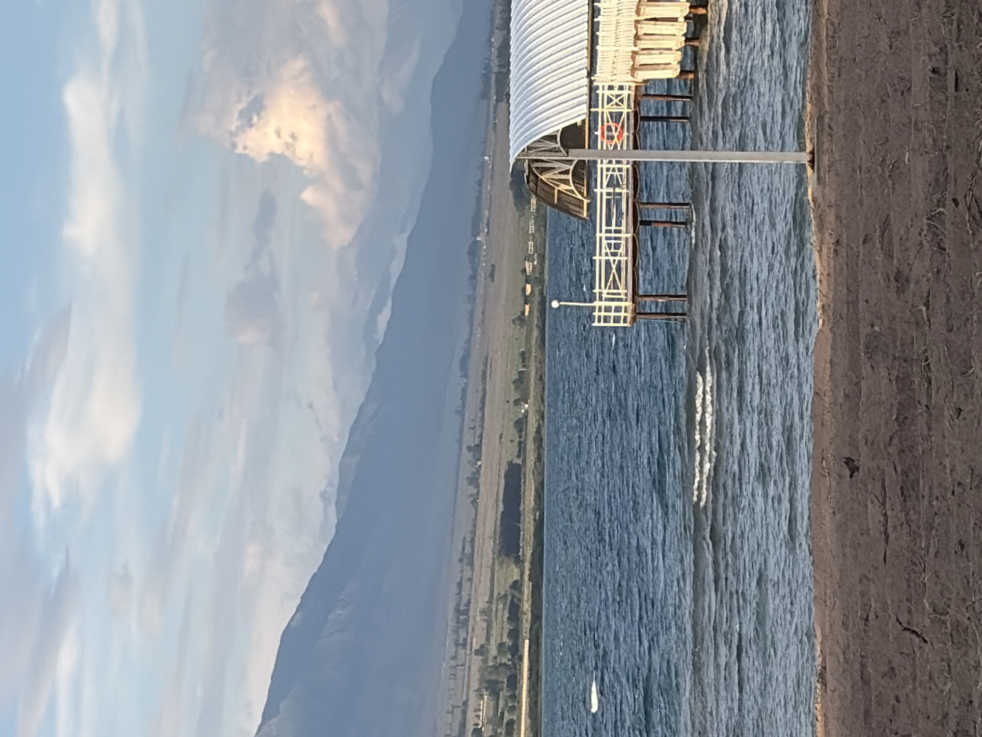 This media file depicts a picturesque outdoor scene, likely taken during the daytime under a cloudy sky. The image is oriented vertically, showing a large body of water, possibly a lake or the sea, with gentle waves lapping against a dark, textured shoreline on the right.
Dominating the right side of the image is a portion of a structure that appears to be a pier or boardwalk. It features a white, corrugated roof and a white railing system. A red life preserver is visible hanging on one of the railings, suggesting a recreational or maritime context. A metal pole supporting the pier extends into the water.
In the background, a long, strip of land stretches horizontally across the frame, featuring a mix of vegetation and scattered structures, possibly indicating a town or settlement along the coast. Beyond this strip of land, a prominent mountain range rises, its slopes shrouded in a hazy, muted blue due to atmospheric perspective or distance. The sky above the mountains is a pale blue, interspersed with dramatic, soft clouds, some of which are illuminated by sunlight, creating a warm glow.
The overall mood of the image is serene and tranquil. The combination of the vast water, the imposing mountains, and the subtle illumination suggests a peaceful, possibly late afternoon or early morning setting, before the full intensity of midday sun. There is no visible text in the image.