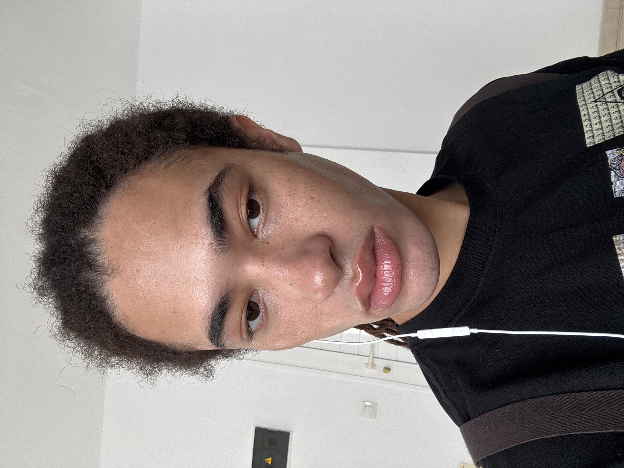 A close-up, tilted, medium-shot photograph presents a person with light skin, brown eyes, and full lips, wearing a black t-shirt. The person's dark, curly hair is short and slightly unkempt. A white earphone cord is visible, suggesting they may be listening to something. The background features a plain white wall, a dark grey panel with a yellow arrow pointing upwards, and a door with brass door hardware. The location is Unknown, Unknown.