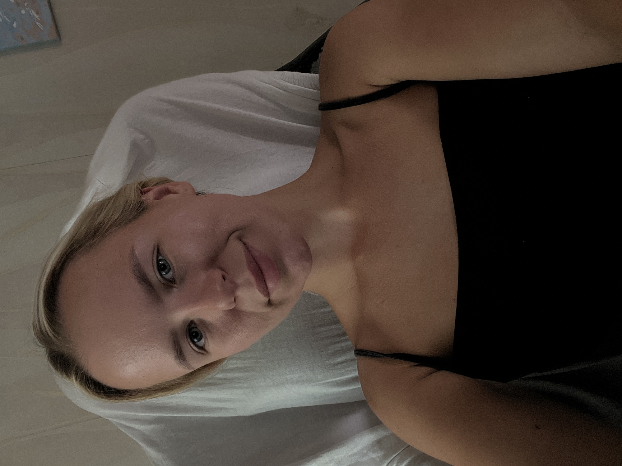 A young Caucasian woman with blonde hair and blue eyes is positioned in the center of the frame, looking directly at the camera. She is wearing a black camisole with thin straps. The background consists of a white sheet draped over a chair and a neutral-colored wall with a small painting. The lighting is soft and ambient, creating a warm glow on her skin. The image appears to be a selfie taken indoors, likely in a bedroom or a similar personal space. The overall mood is relaxed and casual.