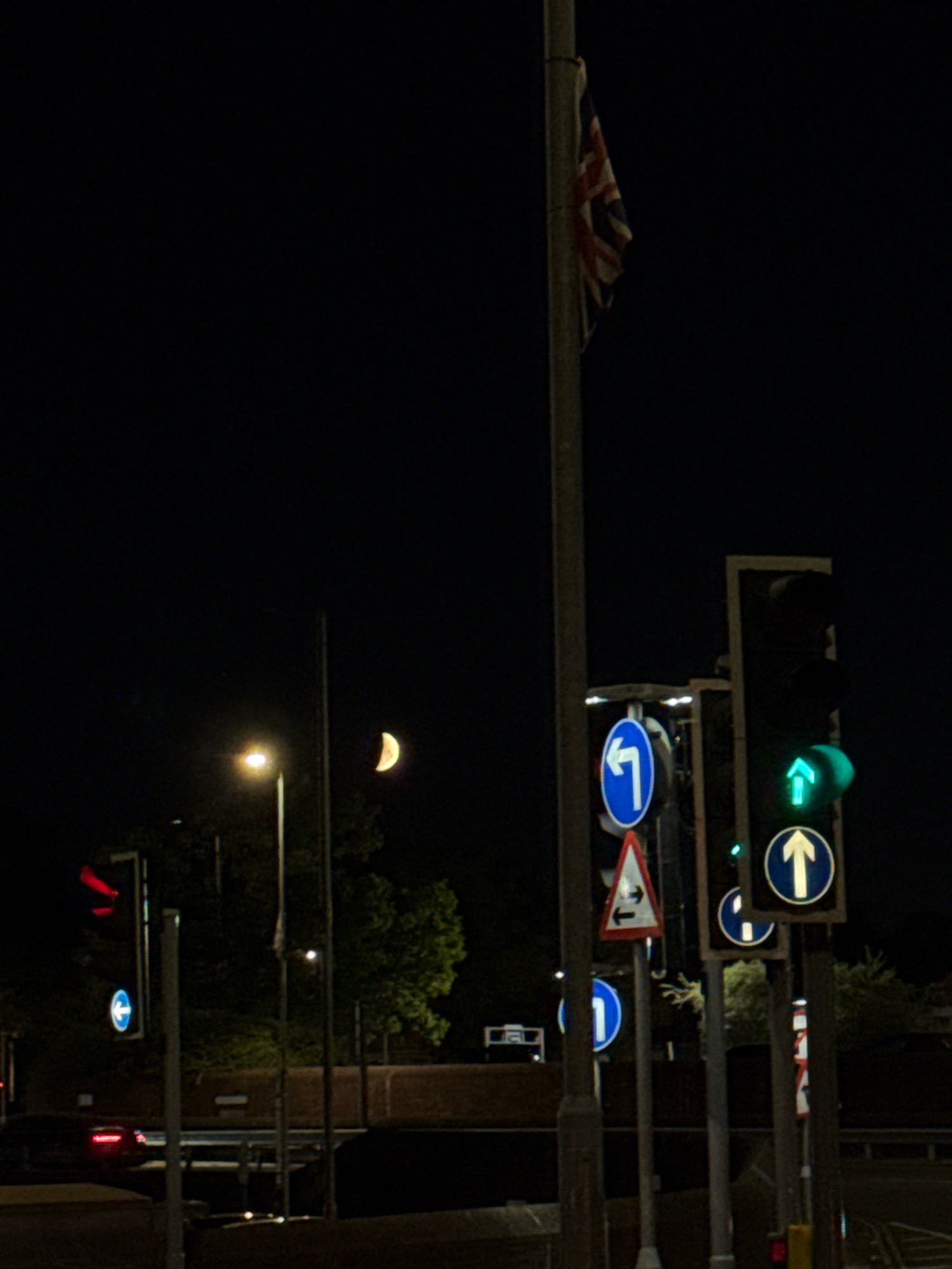 This is a nighttime photograph of a street scene in Basildon, United Kingdom, likely near a junction. A traffic light with a red signal is visible on the left, accompanied by a circular blue sign indicating a mandatory direction. To the right, a traffic light displays a green upward arrow, and below it, a circular blue sign shows a white upward arrow. Another traffic light, largely obscured, also shows a green signal. A British flag is displayed from a flagpole in the background. A crescent moon is visible in the dark sky. Trees and some brickwork are present, suggesting an urban or semi-urban environment. The road and vehicles are dimly lit, indicating a low light condition.