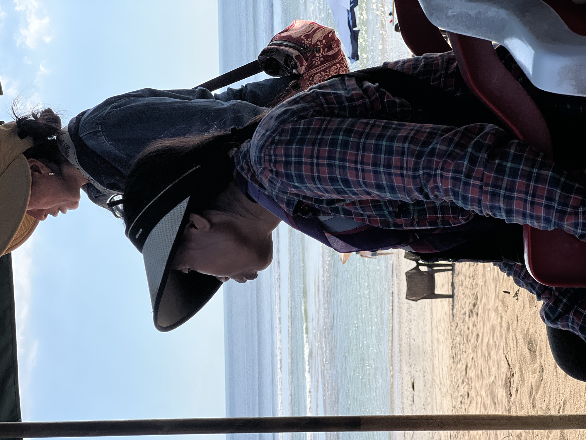 Two individuals, likely tourists, are seated under an awning or shelter on a sandy beach, possibly in Kuta, Indonesia. The person in the foreground is wearing a plaid shirt and a wide-brimmed sun visor, facing to the left, with a slight pout. Behind them, another person, wearing a denim jacket and a sun hat, is partially visible, looking towards the right. Both are seated on chairs, suggesting they are relaxing or observing the surroundings. The background shows a calm ocean with gentle waves, a sandy beach, and two empty chairs. The sky is bright and clear, with a few wispy clouds. The overall scene depicts a casual, seaside setting.