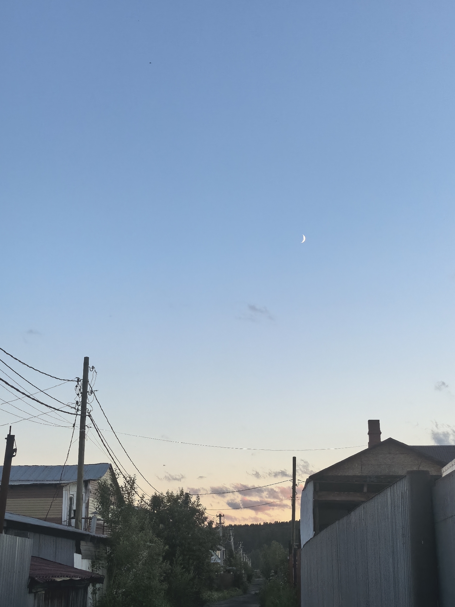 The image depicts an outdoor scene in an urban or semi-urban environment during twilight. The sky is a clear, pale blue, transitioning to lighter hues near the horizon, with a crescent moon visible in the upper right quadrant. A few wisps of clouds are scattered across the sky. In the foreground, the scene is dominated by structures and utility poles.
On the left side of the image, there are several buildings and utility poles with a dense network of overhead wires. One building appears to be made of wood with a metal roof. Further into the scene, a road or path is visible, flanked by vegetation and more buildings in the distance.
On the right side of the image, a corrugated metal fence runs along the foreground, obscuring part of the view. Behind the fence, a building with a prominent brick chimney is visible, partially under construction or in disrepair, with exposed wooden beams.
The overall impression is one of a somewhat weathered, possibly rural or industrial, area at the end of the day. There are no people or animals present in the image.