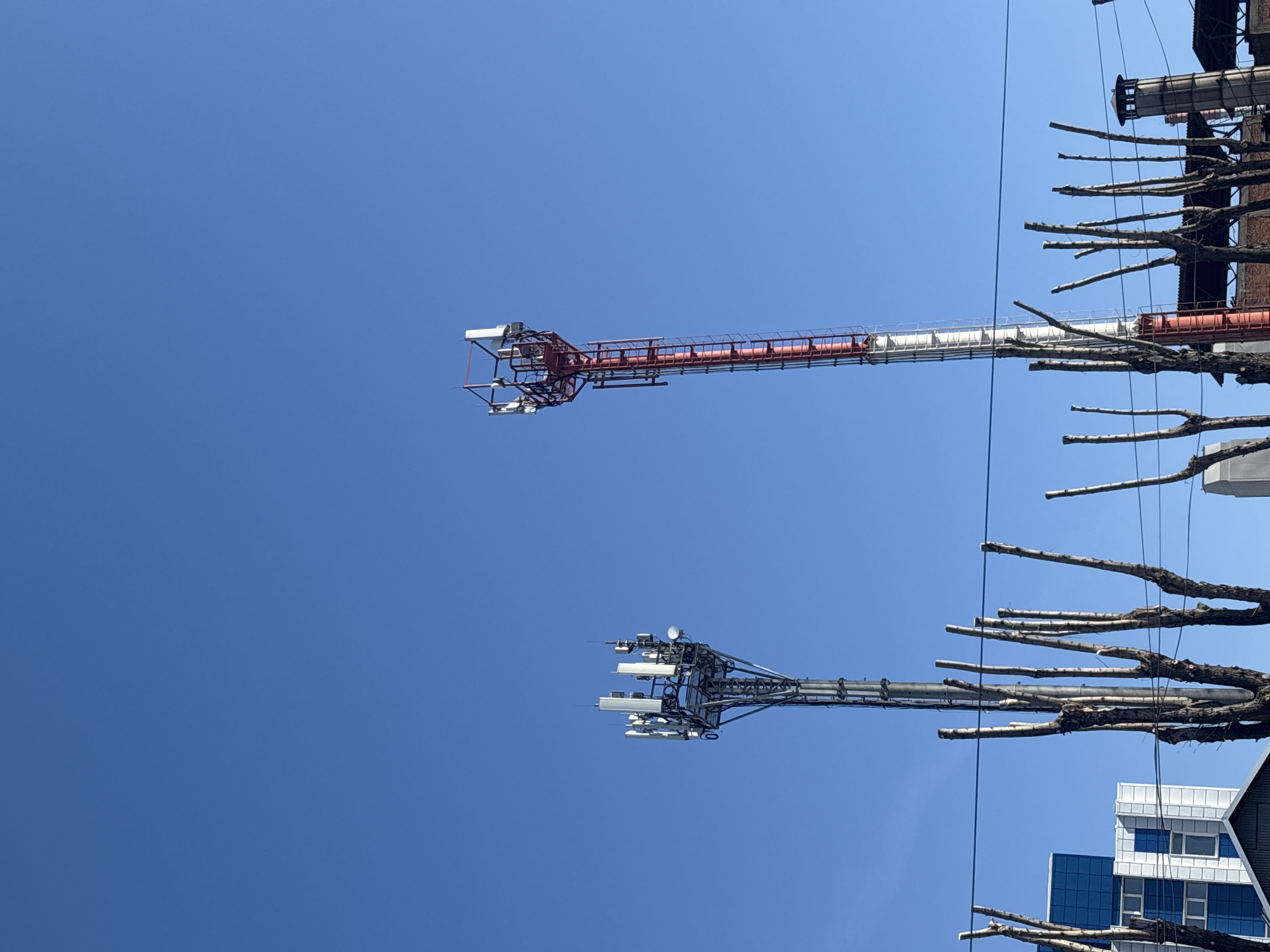 The image depicts two horizontal structures extending into a clear blue sky, partially obscured by tree branches in the foreground. The upper structure is red and white, appearing to be a bridge or crane element. The lower structure is gray and supports communication equipment with multiple antenna-like components. In the bottom right corner of the image, there is a portion of a building with blue and white panels. The location in Perm, Russia is not discernible based on the image alone.