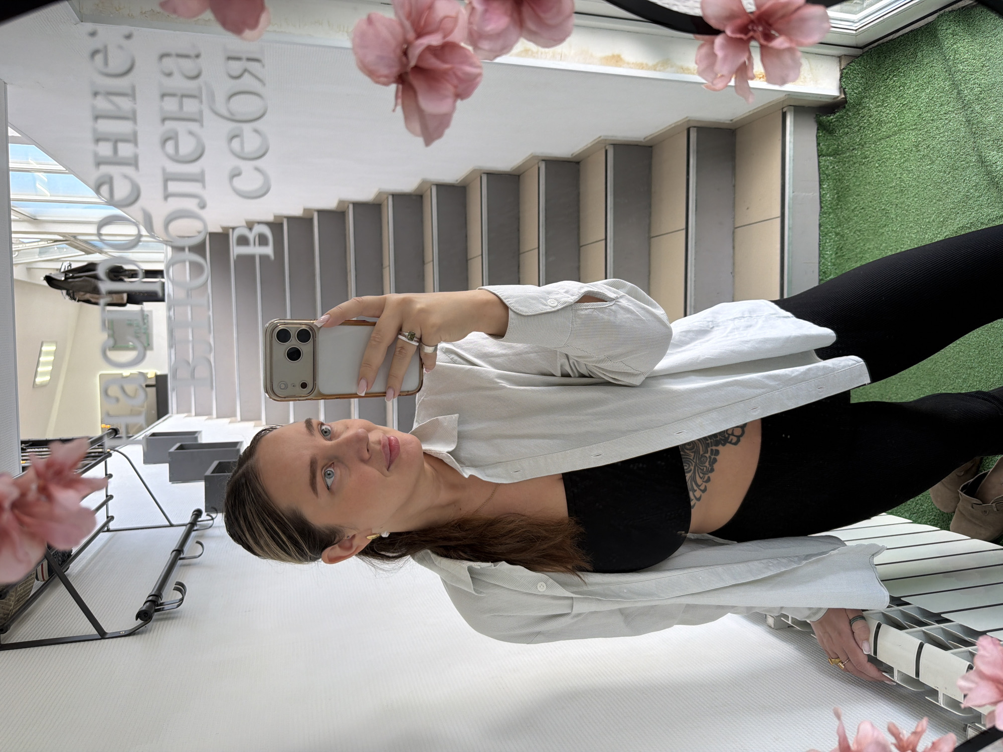 A woman is taking a selfie in a mirror. She is wearing black leggings, a black bra top, and a white button-down shirt. She has light brown hair with blonde highlights. She is holding a silver iPhone. Her tattoo is visible on her side. The background of the mirror shows stairs, a window with pink flowers, and a grassy area. The reflection also shows Russian writing. The location is Chaikovsky, Russia.