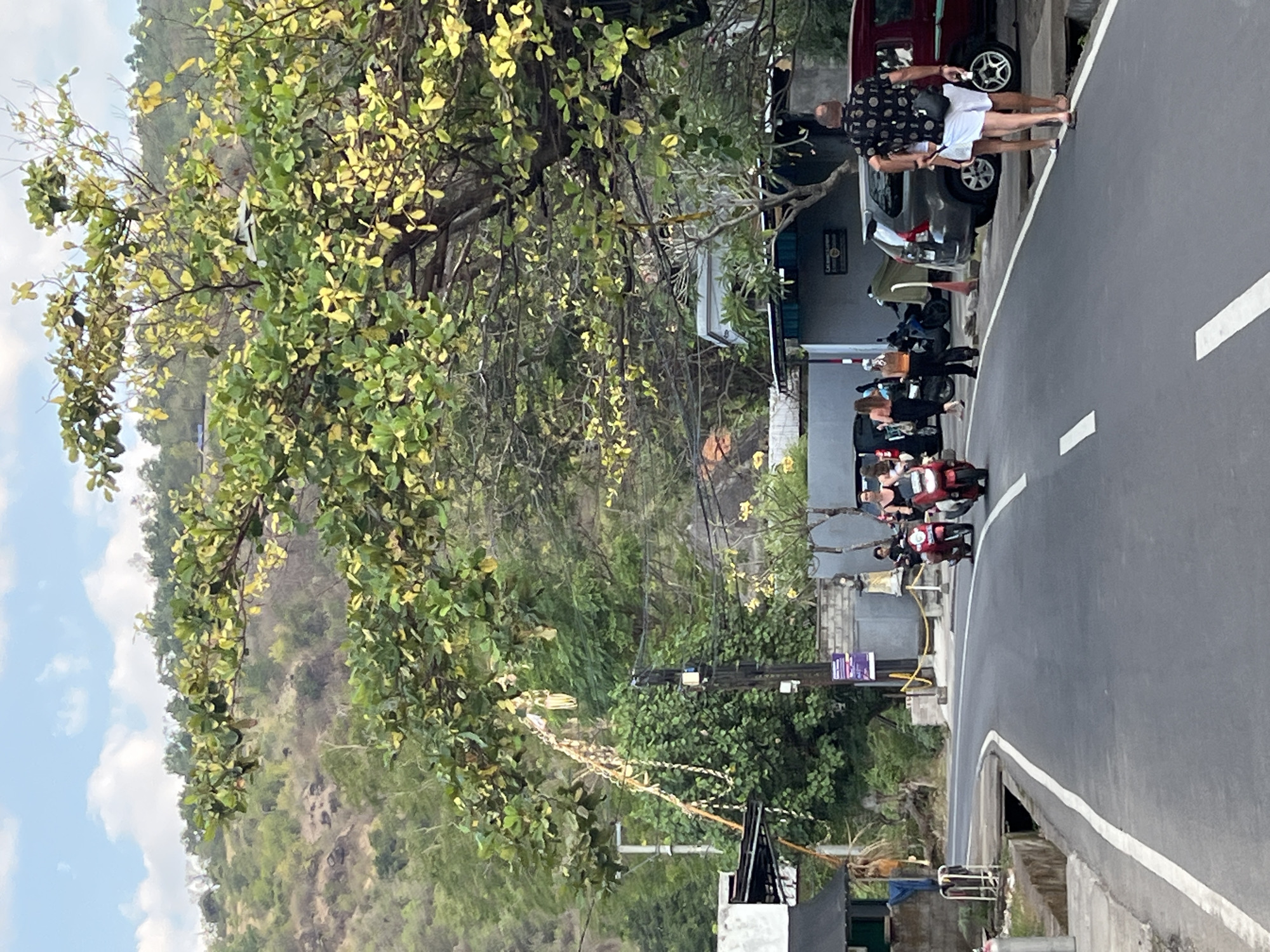 The image depicts a street scene in Abang, Indonesia. A paved road curves to the left, with white lines marking the lanes. On the left side of the road, there are buildings and some vegetation. A man in a black patterned shirt and white shorts is walking away from the camera on the right side of the road, accompanied by a woman in white shorts. Several people are on scooters in the middle of the road. Two red scooters are prominent, one with a rider and a passenger, and another with a single rider. A third scooter, dark blue, is parked on the side. In the background, there are more buildings, power lines, and a lush, green hillside with trees. A large tree with yellowing leaves extends over the road from the right. The sky is partly cloudy.