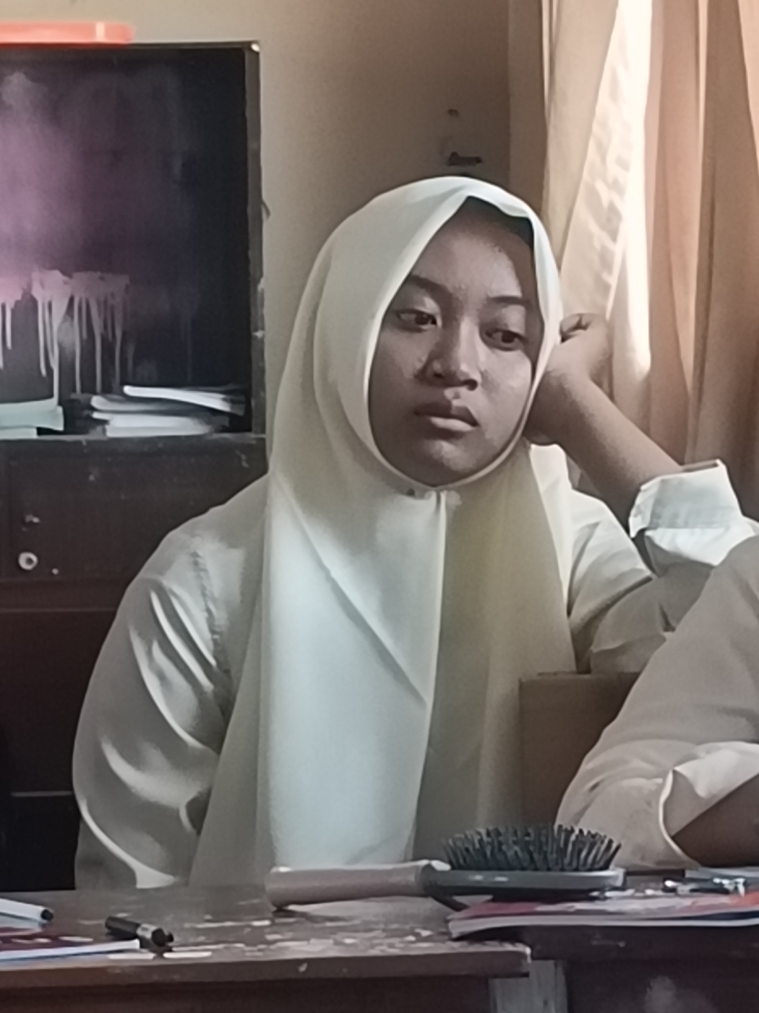 A young Indonesian female wearing a light-colored hijab and shirt is captured in a medium shot, resting her head on her hand and looking off to the side. She is seated at a wooden table in what appears to be a classroom or study environment. On the table, a hairbrush, pens, and papers are visible. In the background, a dark cupboard with some papers stacked on it and a window covered by light brown curtains can be seen. The lighting suggests an indoor setting, possibly during daytime. The scene does not provide specific indicators of its exact location within Kediri City, Indonesia, beyond the general cultural context suggested by the subject's attire.