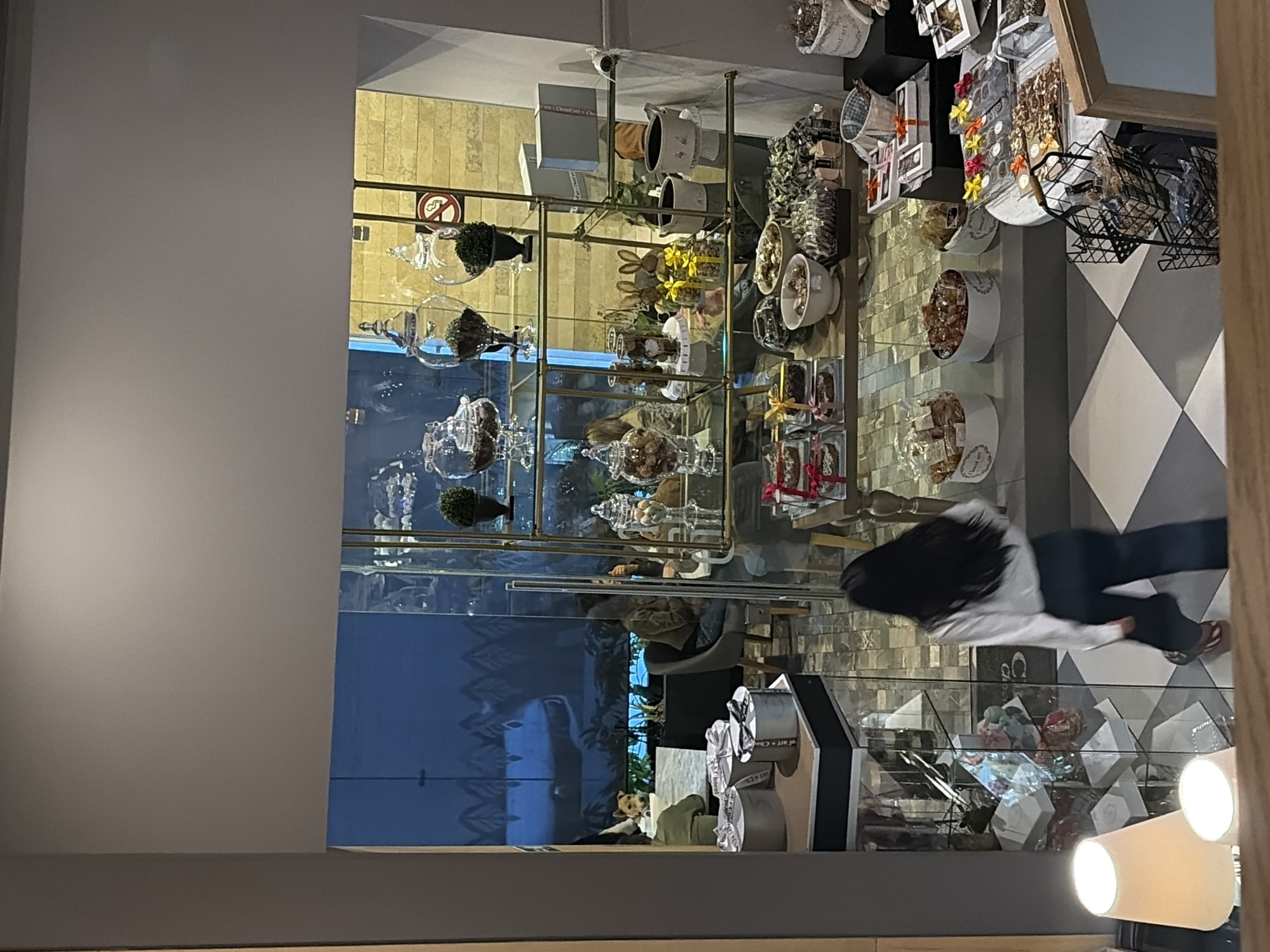 The image displays the interior of a chocolate shop or dessert café, located in the city of Puebla, Mexico, viewed from a slightly elevated perspective.
In the immediate foreground, a female subject with long, dark hair, wearing a white long-sleeved shirt and dark pants, walks from left to right across a floor tiled with a light and dark grey checkered pattern. Two illuminated table lamps are partially visible in the bottom-left corner.
The central section of the establishment features a multi-tiered retail display unit constructed of gold-toned metal and clear glass shelves. These shelves hold various clear glass jars containing confections such as chocolates, nuts, and decorative elements like small plants. Additional merchandise includes packaged chocolates, gift-wrapped boxes (silver and white, some with ribbons), ceramic rabbit figurines adorned with yellow bows, and other small potted plants. A "no smoking" sign is visible on a light-colored, textured wall behind the upper shelves. Lower display surfaces present white circular containers marked "Chocol'art" filled with various sweets, along with bowls and trays of individually wrapped items. A white, round display table on the right holds more packaged confectionery, accompanied by a black wire basket display unit.
Beyond the main retail area, separated by a clear glass partition, is a seating section. At least three individuals are visible, seated at tables with modern chairs. On the left, a person wearing a green top is partially seen, seated across from a small, light-brown dog positioned on a chair, looking towards the camera. Further back, another person in a brown jacket is seated. The background of the seating area features a dark blue mural with a stylized pattern, and a large planter containing dark foliage is present.
The overall environment is well-lit by a combination of ambient and overhead lighting fixtures.