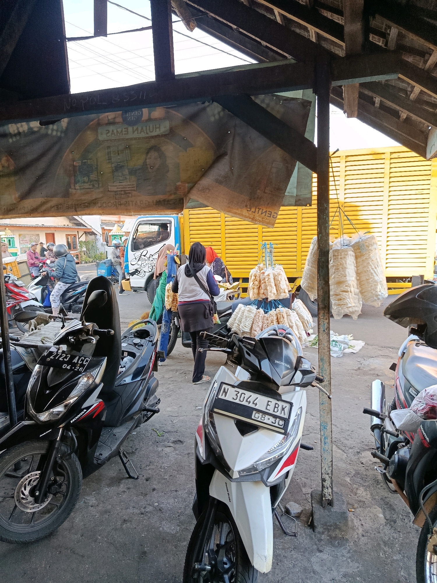 A street market scene unfolds in Bululawang, Indonesia, under a rustic, wooden-beamed shelter. The ground is a mix of concrete and dirt, with several motorcycles parked. In the foreground, a white Honda Vario motorcycle with license plate N 3044 EBK is prominent. To its left is a black Honda motorcycle with license plate W 2723 ACC.
Behind the motorcycles, a blue and white truck is parked, its rear portion visible. The truck is adorned with a stylized drawing. To the right of the truck, large bunches of what appear to be fried snacks, possibly crackers or chips, are hanging from strings, wrapped in plastic.
Several individuals are present in the scene. A woman in a black hijab and grey top is seen walking away from the camera. Another woman in a pink headscarf is visible near the truck. A man in a blue shirt and helmet is standing by the truck. Other people are seen in the background, some on motorcycles.
A large banner hangs from the shelter's beams, displaying text in Indonesian, some of which is reversed. The backdrop consists of buildings and a bright yellow structure with horizontal slats, possibly a shuttered wall. The lighting suggests it is daytime, with sunlight filtering through the shelter and illuminating parts of the scene.