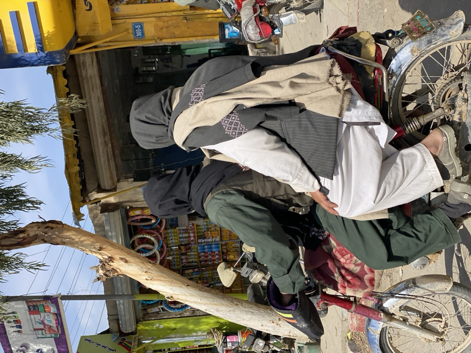 A daytime street scene in Kandahar, Afghanistan, depicts two individuals seated on a red motorcycle positioned in front of a row of commercial establishments. The driver, seated forward, wears dark green long-sleeved clothing, a dark vest, and a black head covering. Their hands are on the motorcycle's handlebars, encased in dark padded gloves with orange, yellow, and red stripe detailing. Directly behind the driver, a second individual is seated, wearing white traditional clothing (shalwar kameez), a dark grey pinstriped vest, and a light beige, patterned shawl draped over their shoulders. This passenger also wears a dark head covering and grey slip-on shoes. A red and white patterned blanket is draped over the motorcycle's seat.
To the left, a shop entrance displays various items, including stacked cans, bottles, and colorful rings, under a white banner with text. A light-barked tree trunk rises from the lower left foreground. To the right, another commercial storefront features a closed corrugated metal roller shutter, a yellow awning, and a blue sign with "305-5" in white numbers. A second motorcycle's rear section is partially visible further down the street on the right. The ground beneath the motorcycle appears to be an unpaved, dusty surface. The sky is bright blue with scattered white clouds.