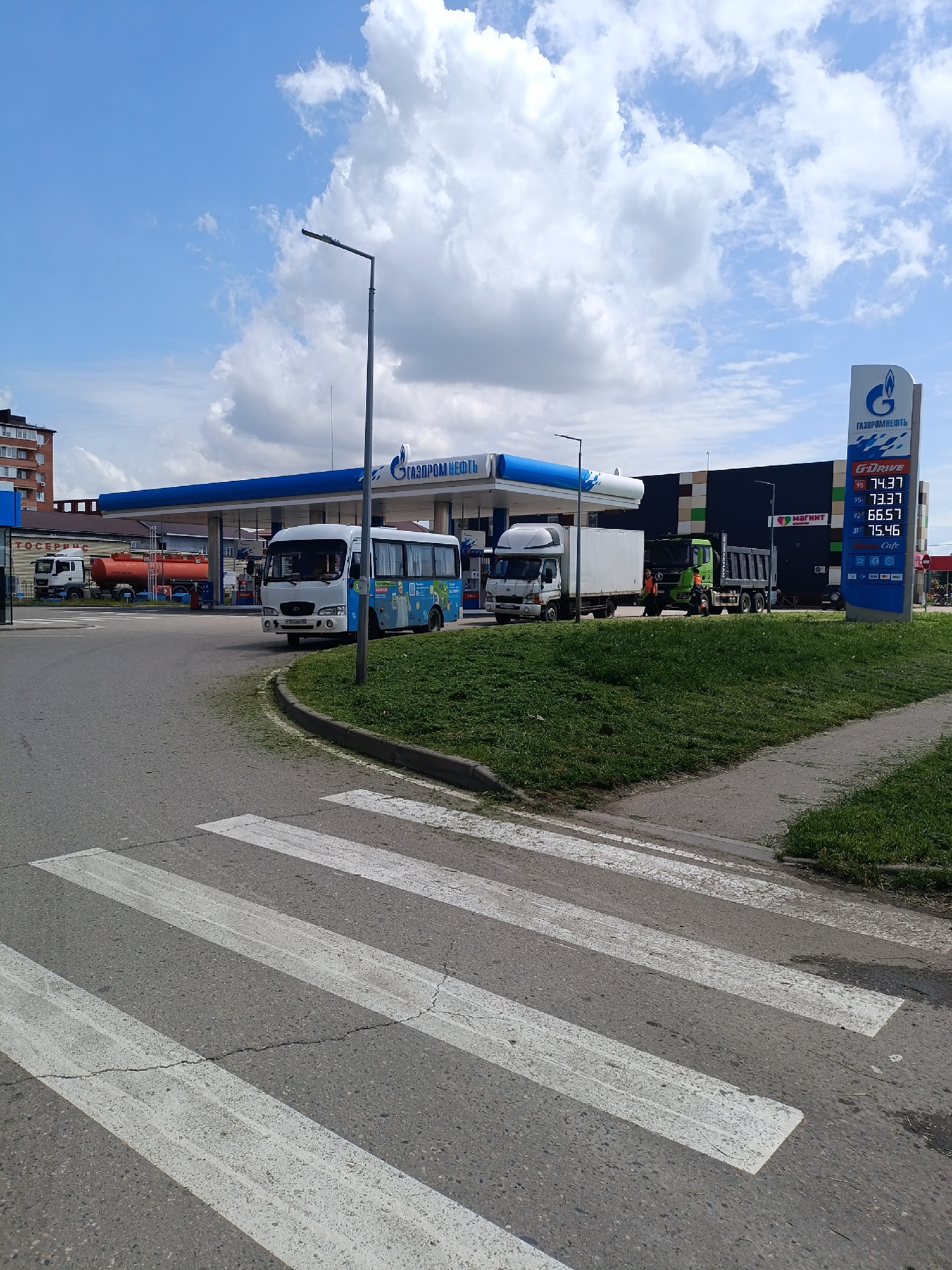 The image captures a street scene in Yablonovskiy, Russia, featuring a Gazpromneft gas station. The gas station has a prominent blue and white logo, a gas price sign listing prices for different types of fuel, and a store with a "Magnet" sign. Several vehicles are visible at the gas station, including a white bus, a white truck, a green truck, and a fuel tanker truck. Two people wearing orange vests are near the green truck. A pedestrian crosswalk is visible in the foreground of the image, with a view of the cloudy blue sky. The surrounding environment includes green grassy areas and a few buildings in the background.