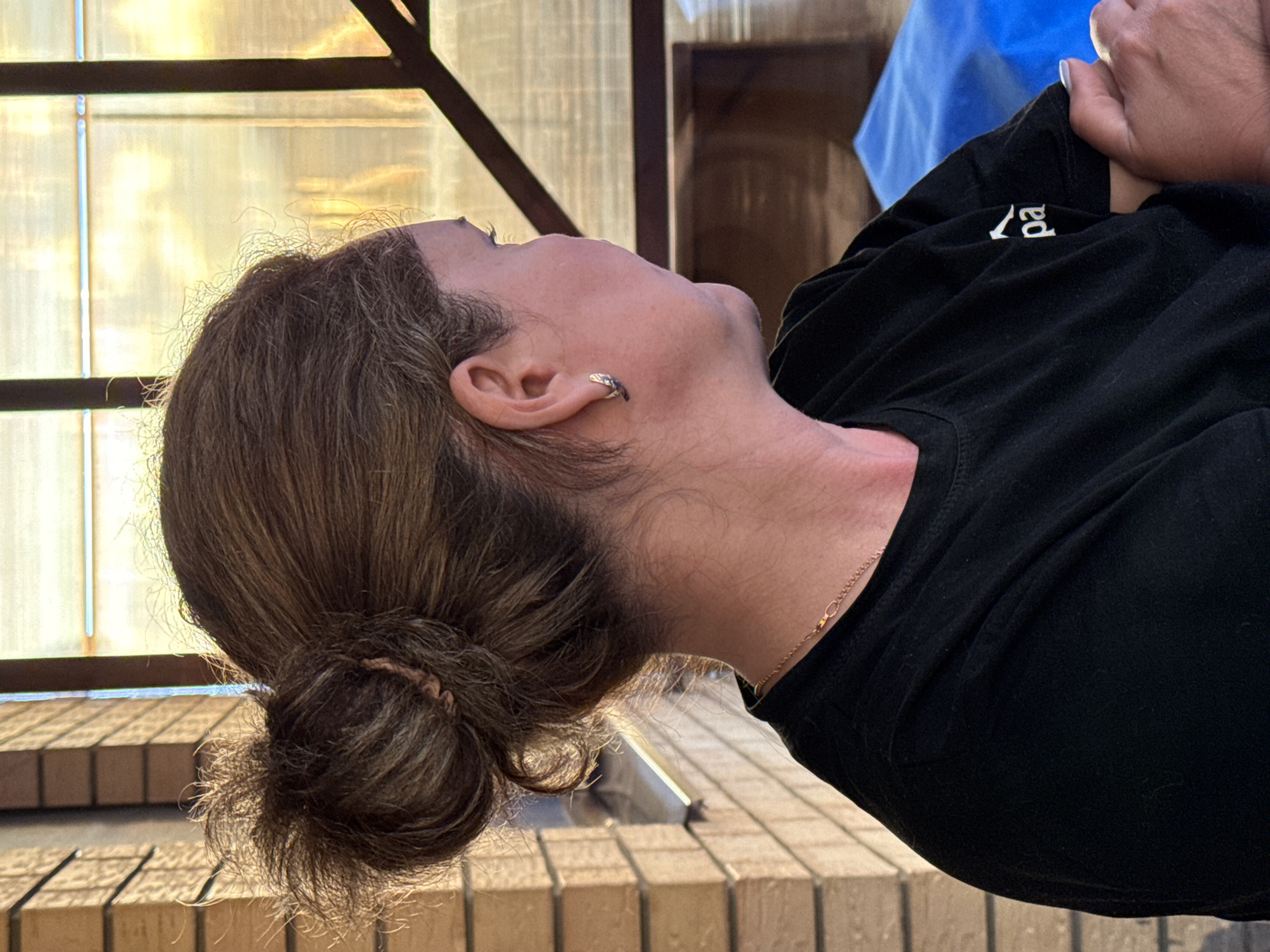 This is a close-up, rotated image of a person's head and upper torso. The person has brown hair pulled back into a bun at the nape of their neck, with some wisps of hair around their forehead and ears. They are wearing a black t-shirt with a white logo on the left side of the chest, which appears to read "ropa". A thin, delicate gold chain necklace is visible around their neck. The person has an earring in their left ear. The background suggests an outdoor setting, possibly a patio or balcony, with wooden structures and a translucent, yellowish material that could be a screen or awning. The lighting suggests it is daytime, with sunlight illuminating the scene and casting shadows. The overall impression is a candid, informal portrait.