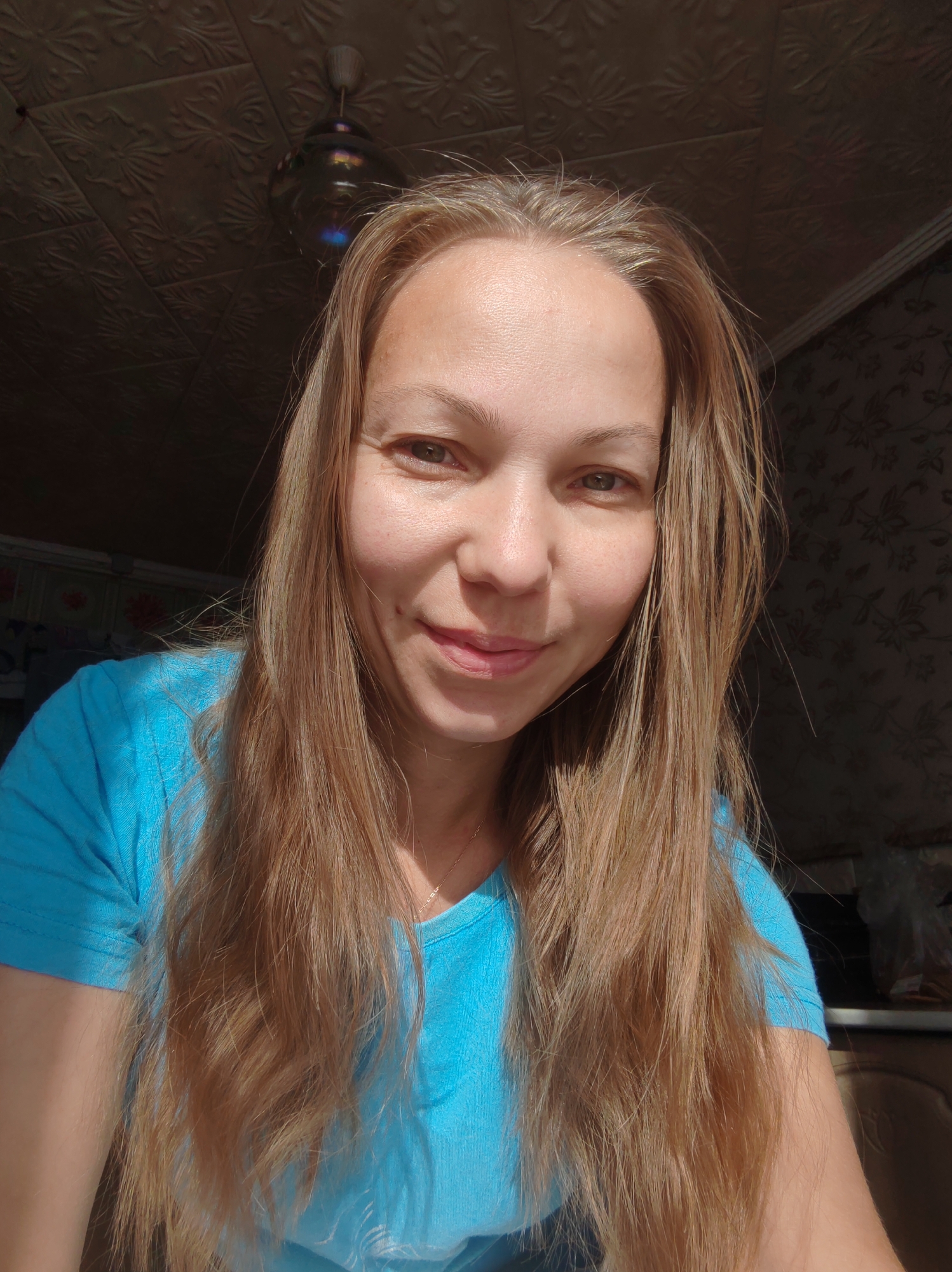 A medium shot shows a woman with long, light brown hair, wearing a blue t-shirt. She is looking towards the camera with a slight smile. Her face is illuminated by natural light, creating shadows on her skin. The background appears to be an indoor setting, with a textured ceiling and a patterned wallpaper visible. A light fixture hangs from the ceiling. The overall impression is of a casual, candid photograph taken indoors.