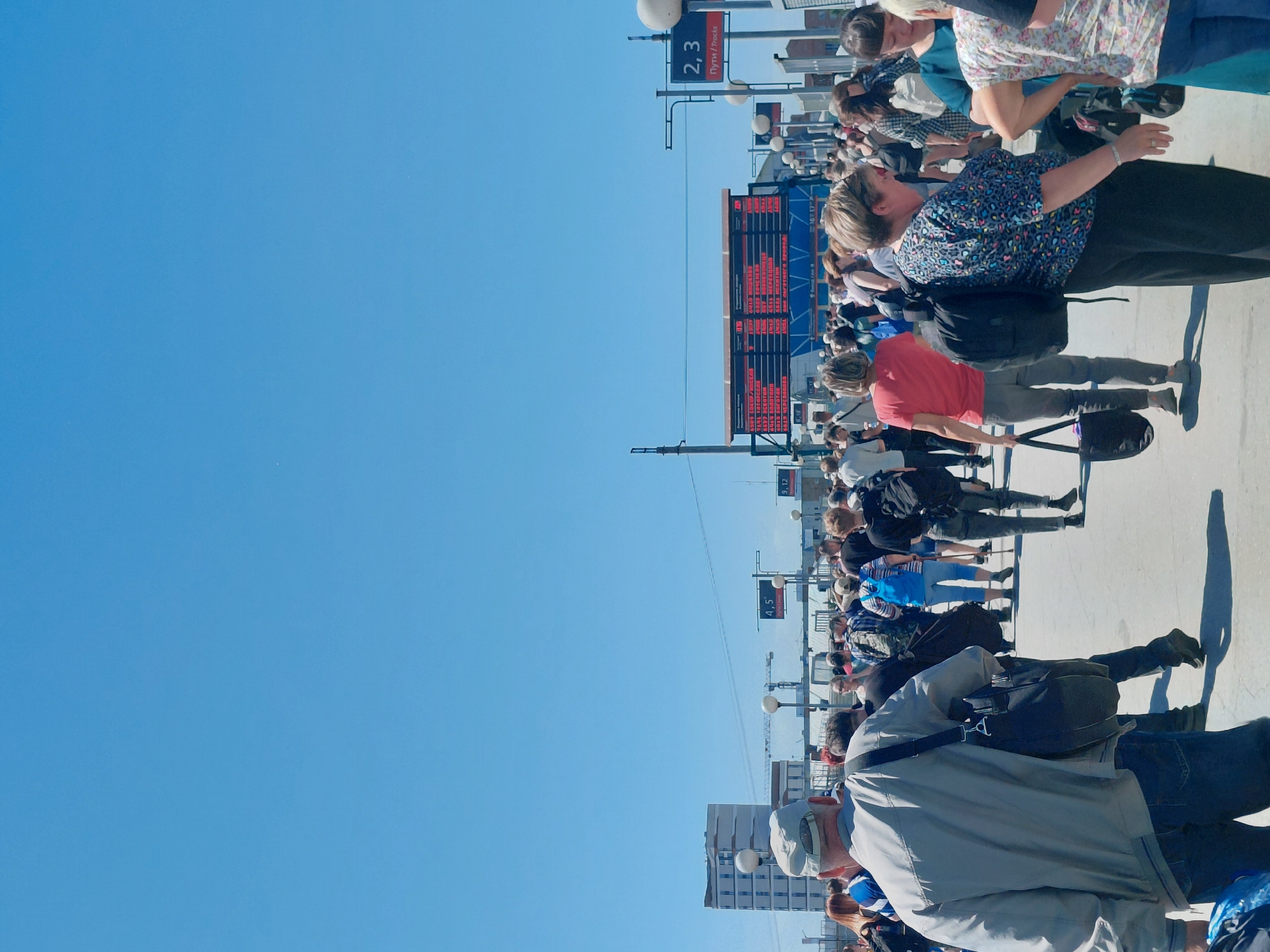 This is a full, eye-level, outdoor shot of a large crowd of people walking in what appears to be a train station or transportation hub under a clear blue sky. In the background, a tall, modern building and construction cranes are visible, suggesting an urban environment. A large digital display board shows train schedules and destinations, with some text in Cyrillic. Several people are carrying luggage and bags, indicating travel. The ground is paved, and the scene is bathed in bright sunlight, casting distinct shadows. The overall atmosphere is one of movement and activity.
